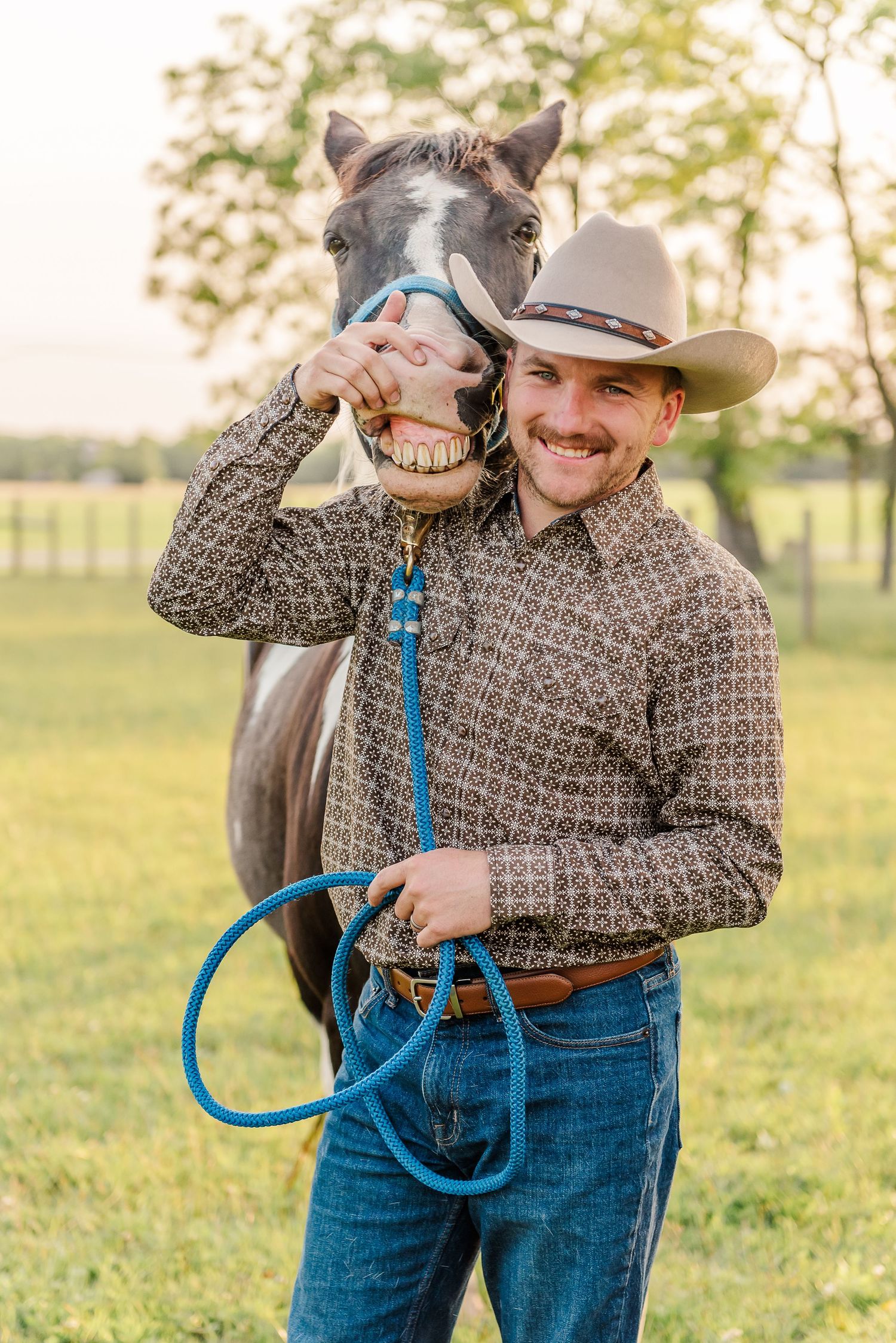 A person in western attire holds a blue rope while posing in a grassy field at sunset.