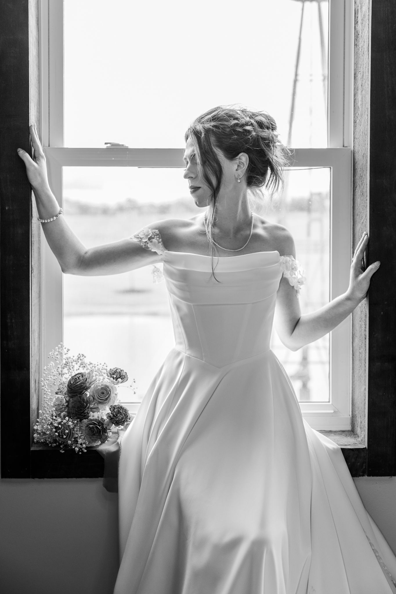 Black and white portrait of a bride in a satin gown standing at a window with bouquet beside her.The Old Barns at Dry Run Farms in Ohio