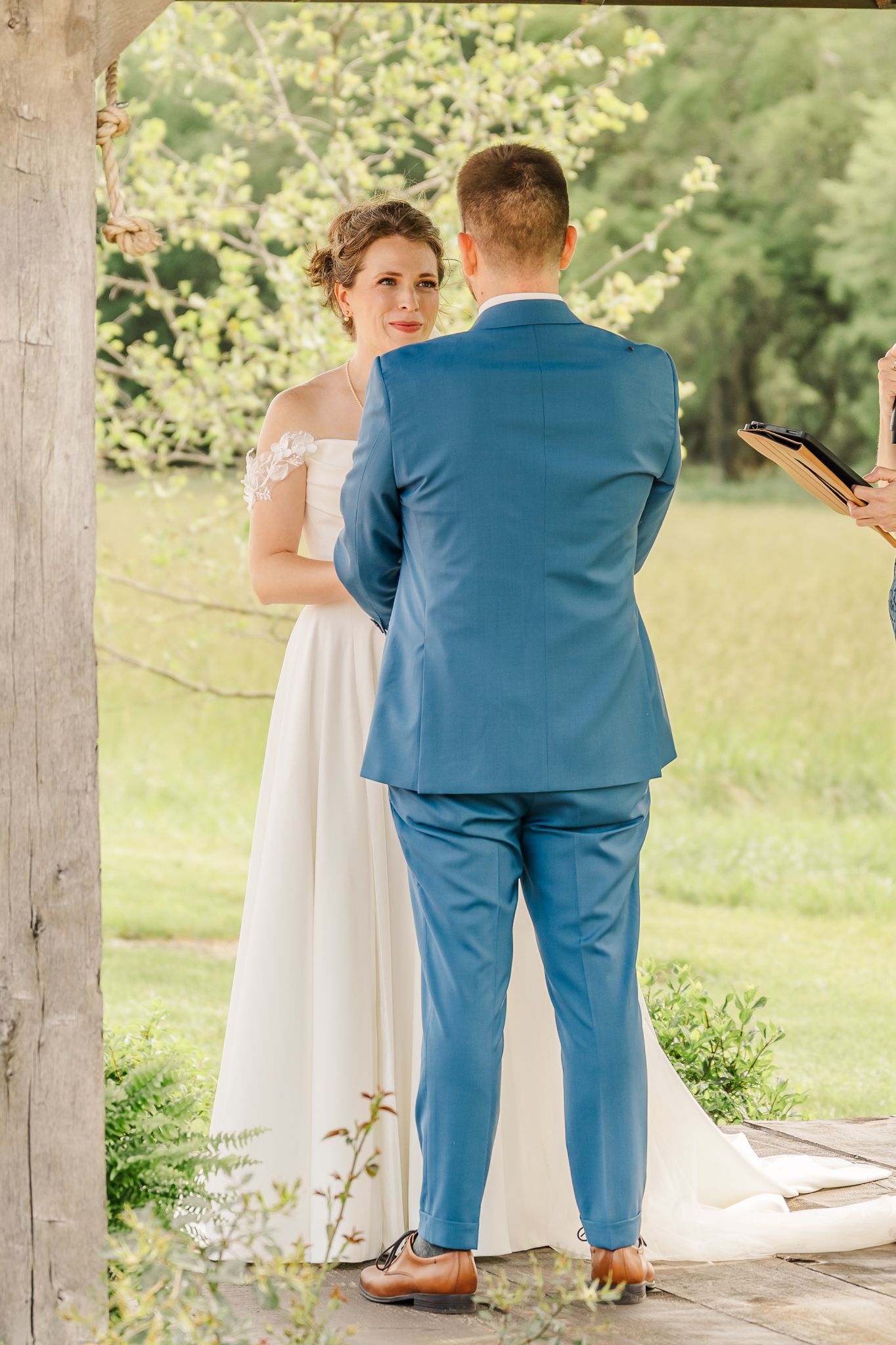 Bride in white gown & groom in blue suit exchange vows outdoors at a garden wedding ceremony.The Old Barns at Dry Run Farms in Williamsport.