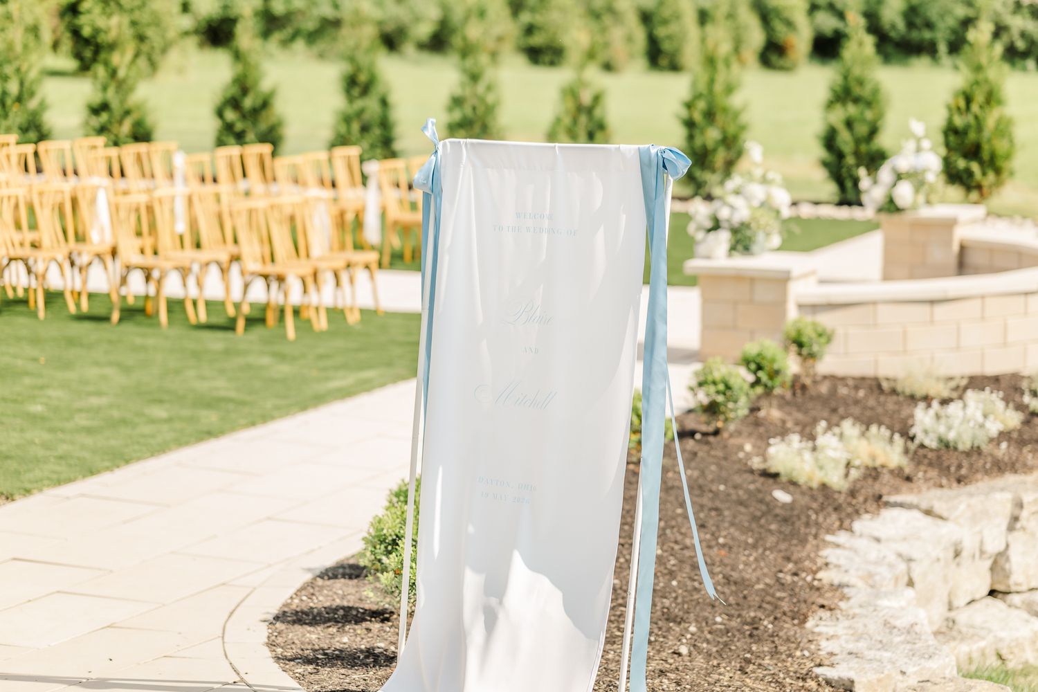 Outdoor wedding ceremony setup with white fabric aisle marker, wooden chairs on green lawn, & landscaped garden backdrop. Canopy Creek Farm