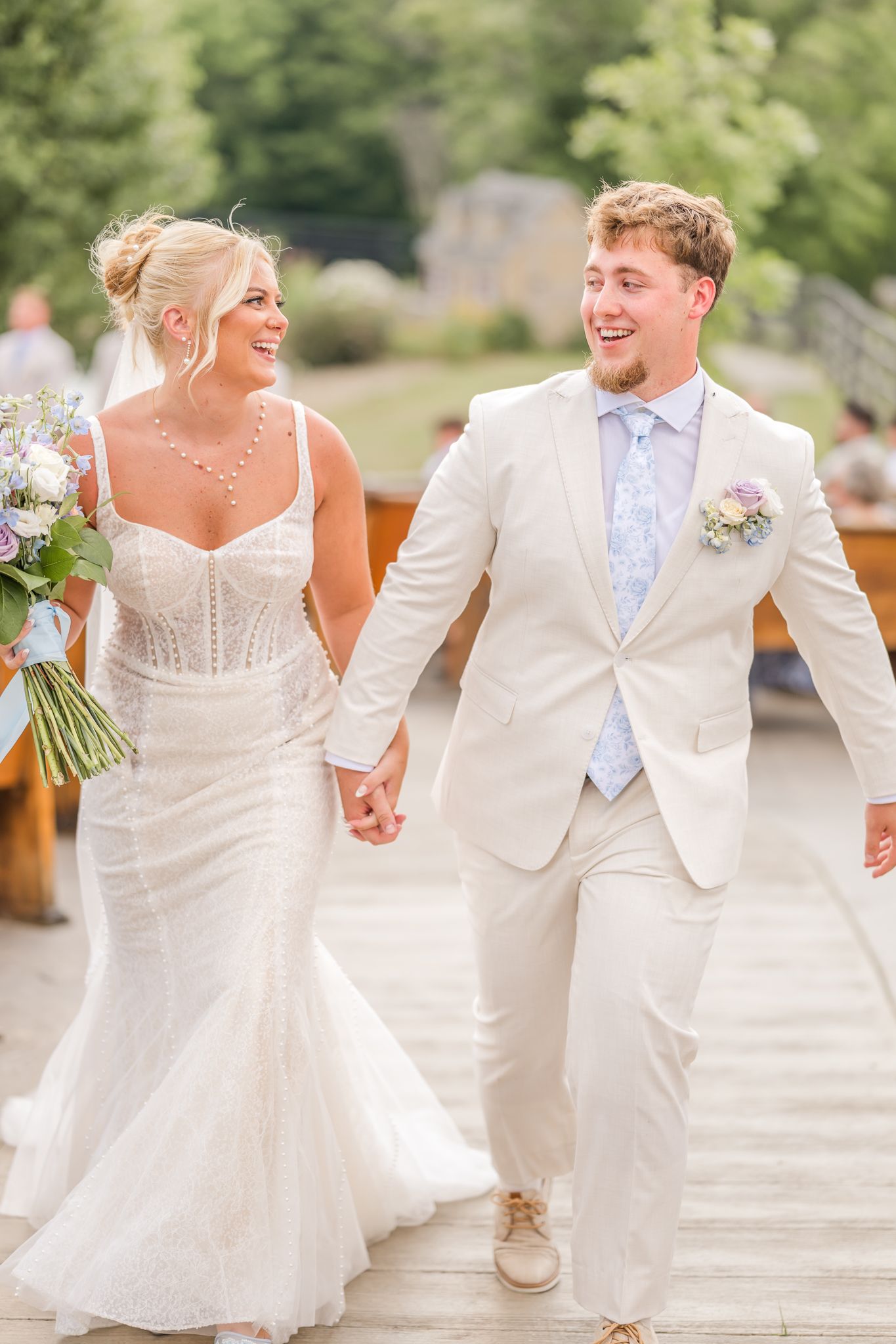 Bride and groom walking hand in hand, smiling joyfully, bride in white gown holding bouquet, groom in cream suit. Peacock Ridge Venue