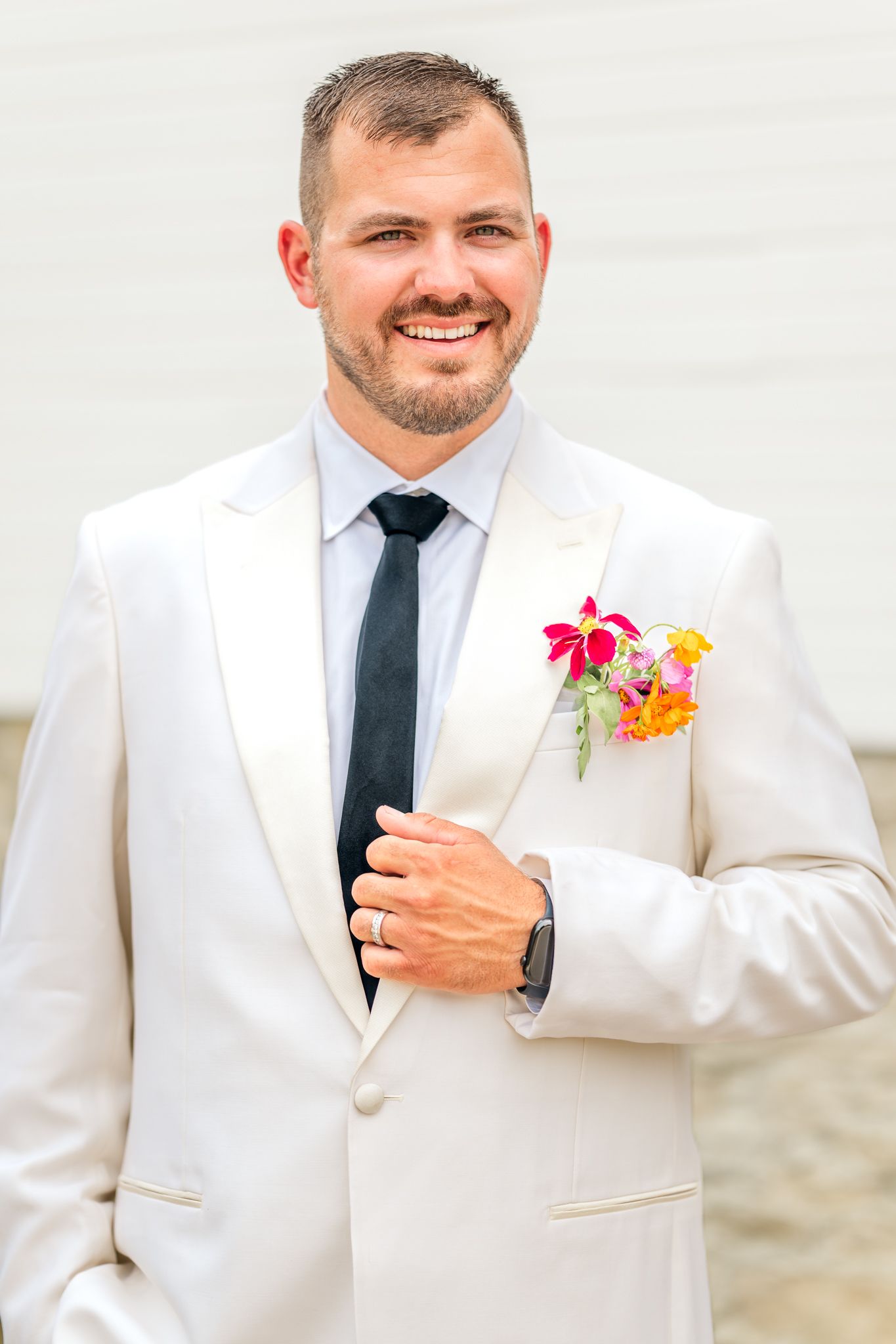 Smiling groom in white tuxedo jacket with black tie and colorful floral boutonniere poses confidently outdoors. The Crawford Barn in Ohio.