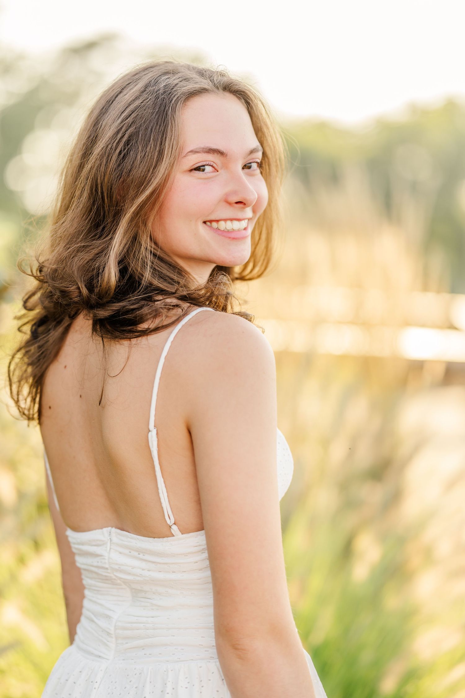 Young woman in white sundress smiling over shoulder outdoors in golden hour sunlight near tall grass.
