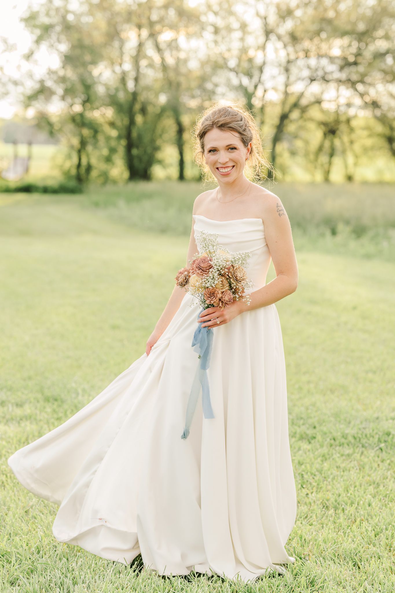 Smiling bride in white strapless gown holding wildflower bouquet with blue ribbon, standing on green grass at golden hour.