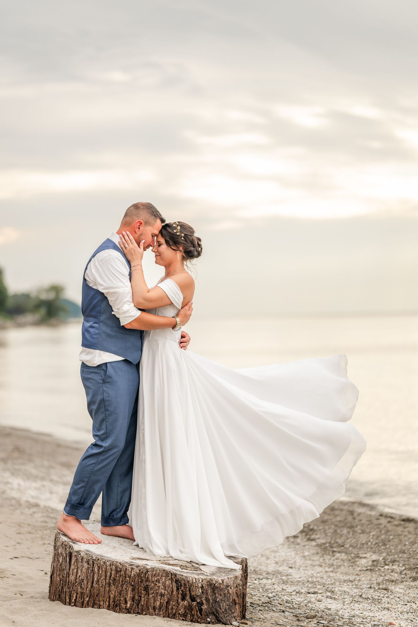 Bride and groom share a romantic beach moment, her white gown flowing in the breeze at sunset. Lake Erie Ohio Wedding.