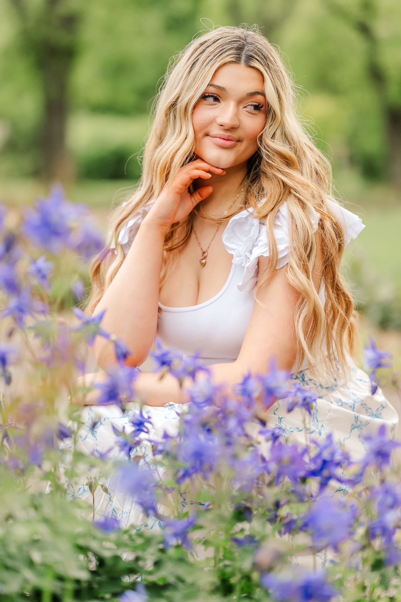 Young woman with long wavy blonde hair in a white ruffled top poses thoughtfully among purple wildflowers in a sunlit green meadow.
