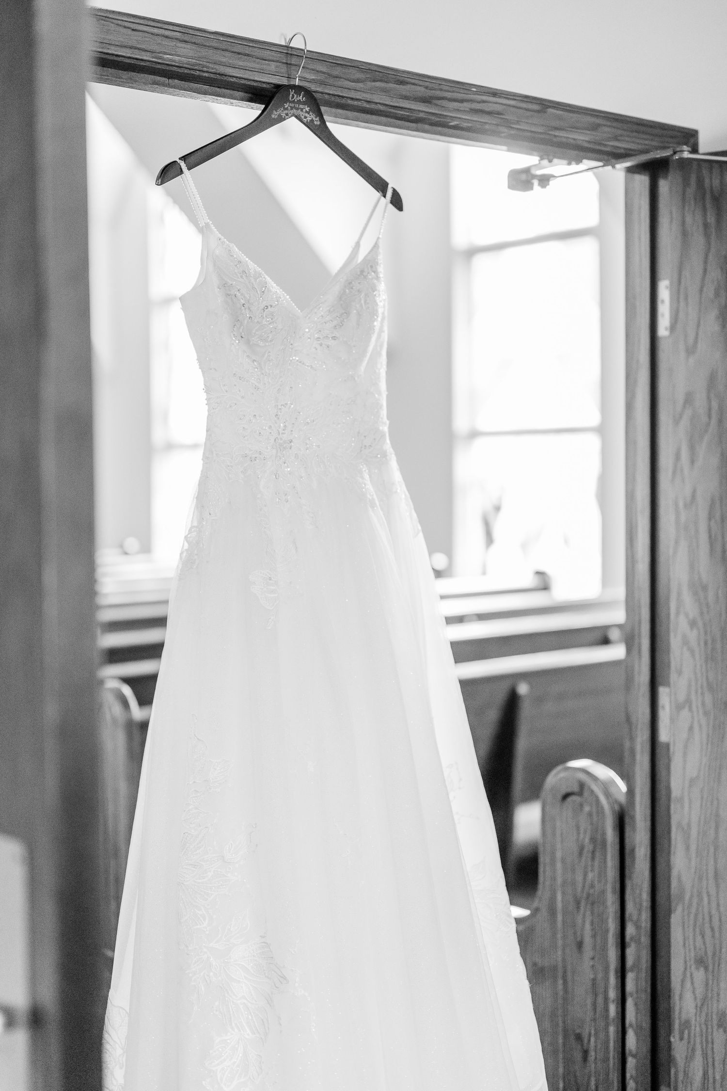 Elegant white wedding dress with lace details hanging by window in black and white photography.