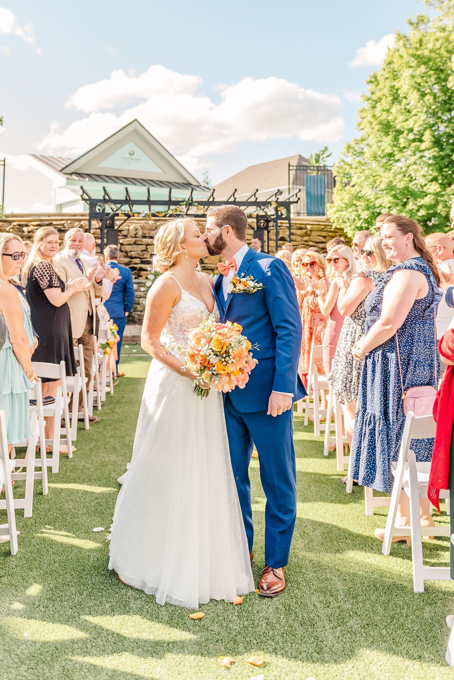 Newlyweds share a kiss surrounded by guests during outdoor wedding ceremony.
