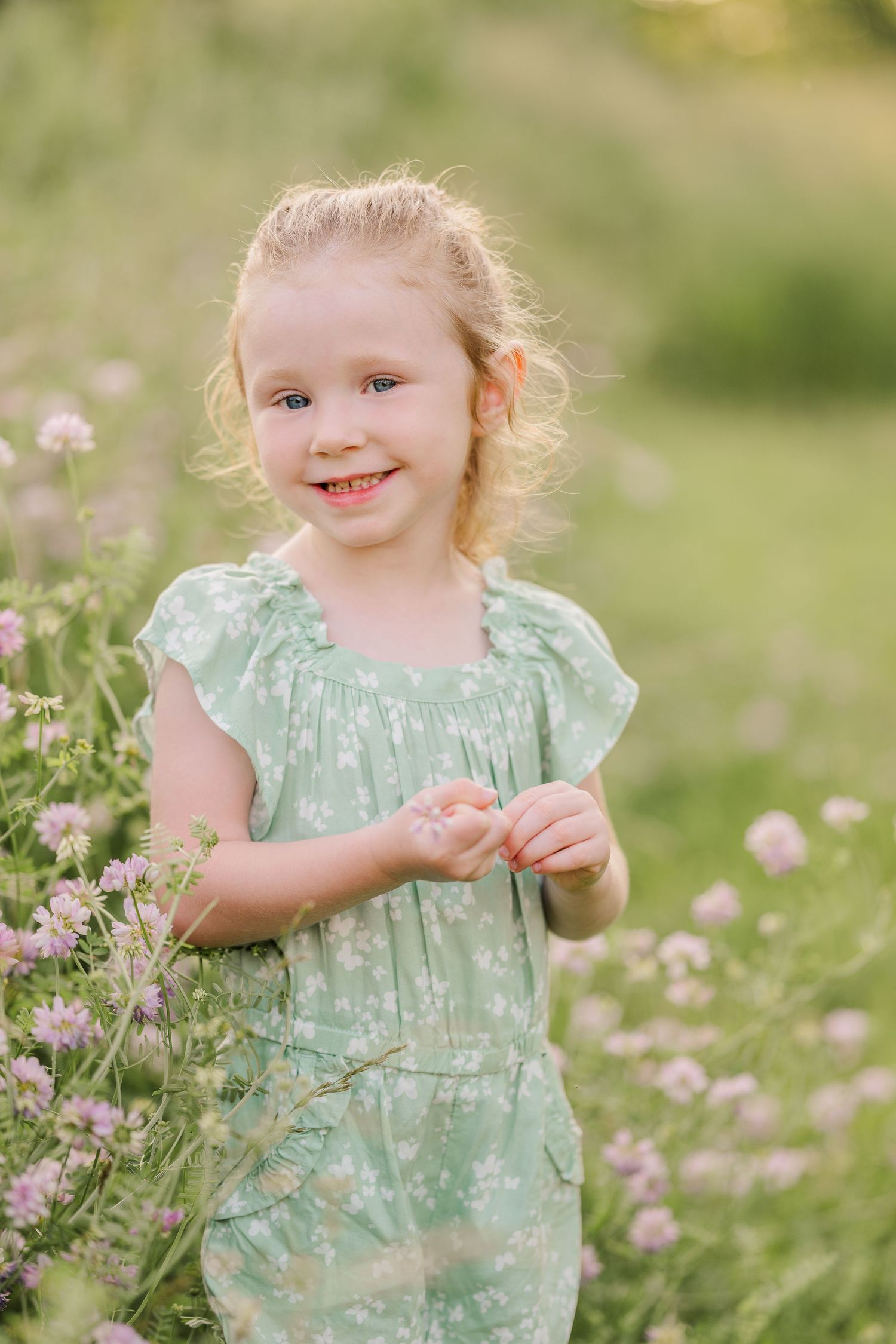 Child in mint green dress plays among pink wildflowers in spring meadow.