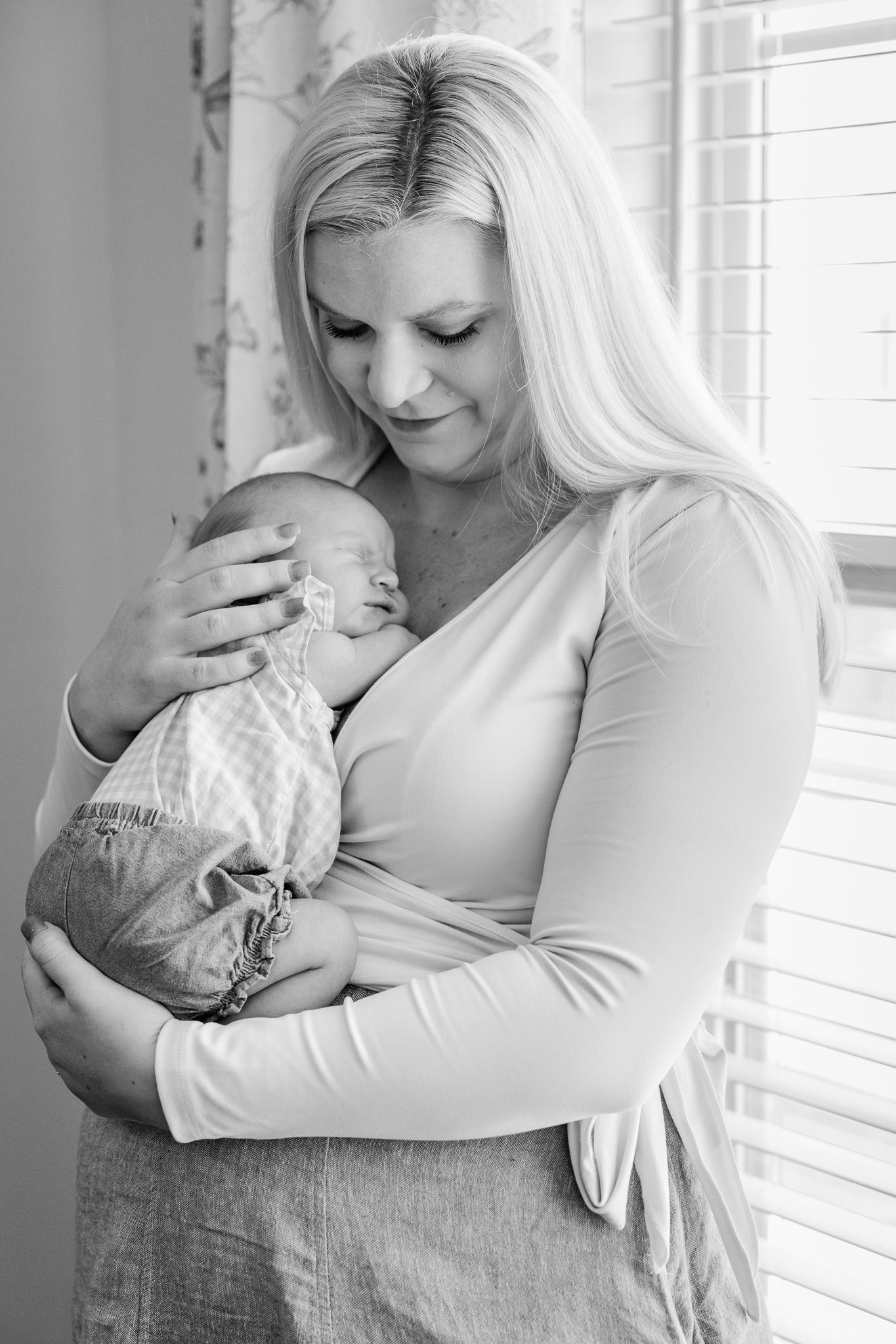 Black and white photo of a smiling blonde mother tenderly cradling her sleeping newborn baby by a window.