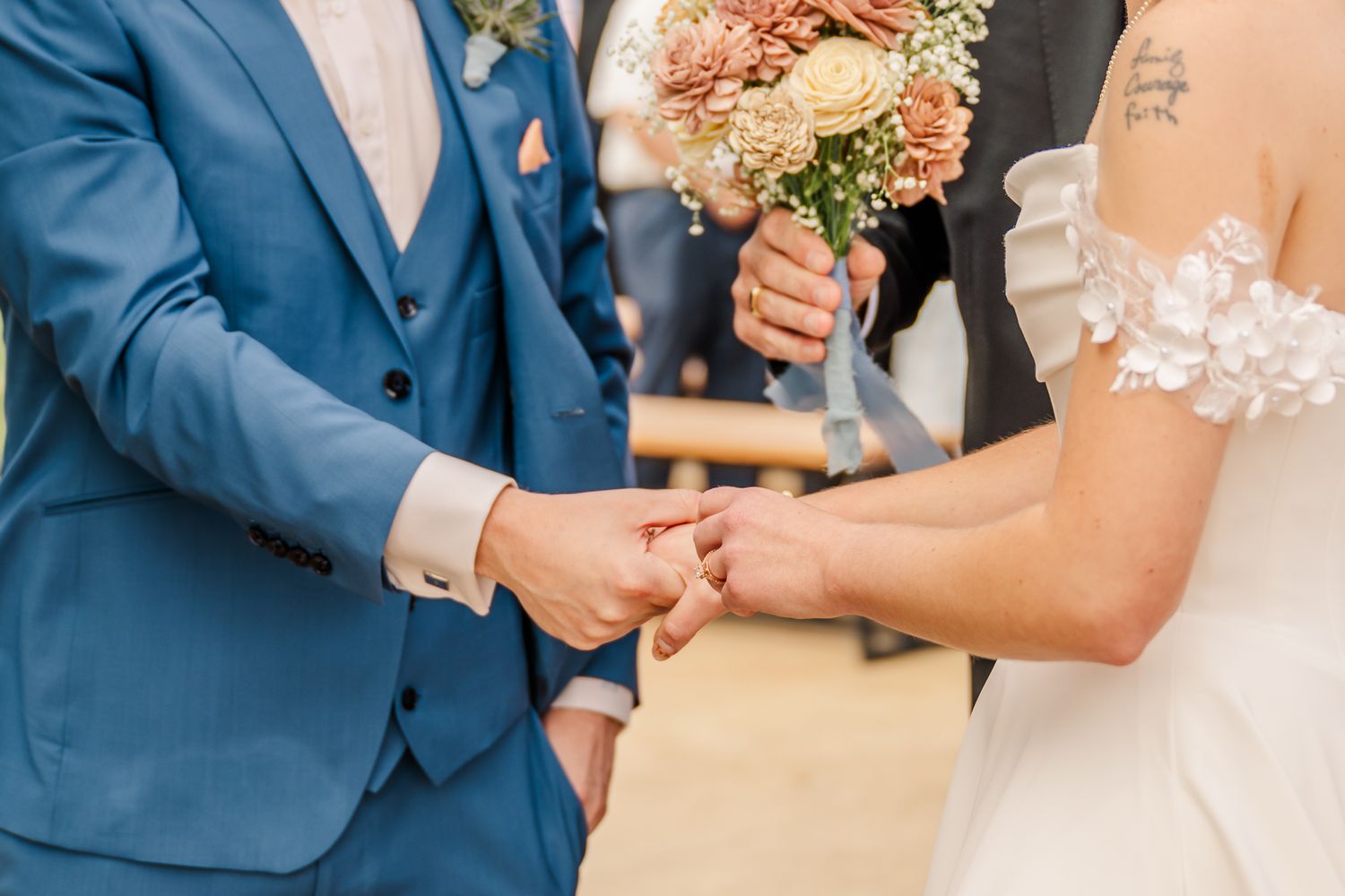 Groom in blue suit placing wedding ring on brides finger during ceremony, bride holding pink floral bouquet.The Old Barns at Dry Run Farms.