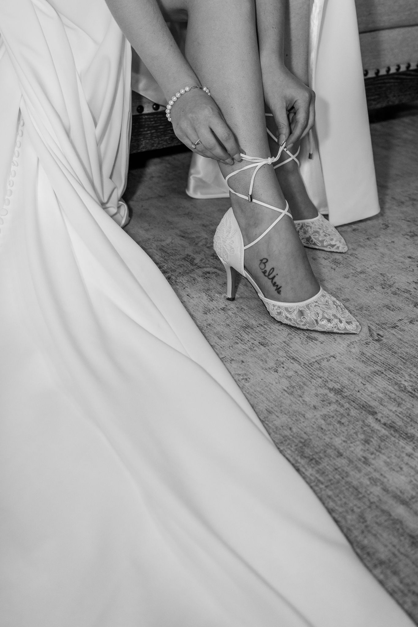 Bride tying lace strappy heels on her wedding day, wearing a flowing white gown and pearl bracelet. The Old Barns at Dry Run Farms.