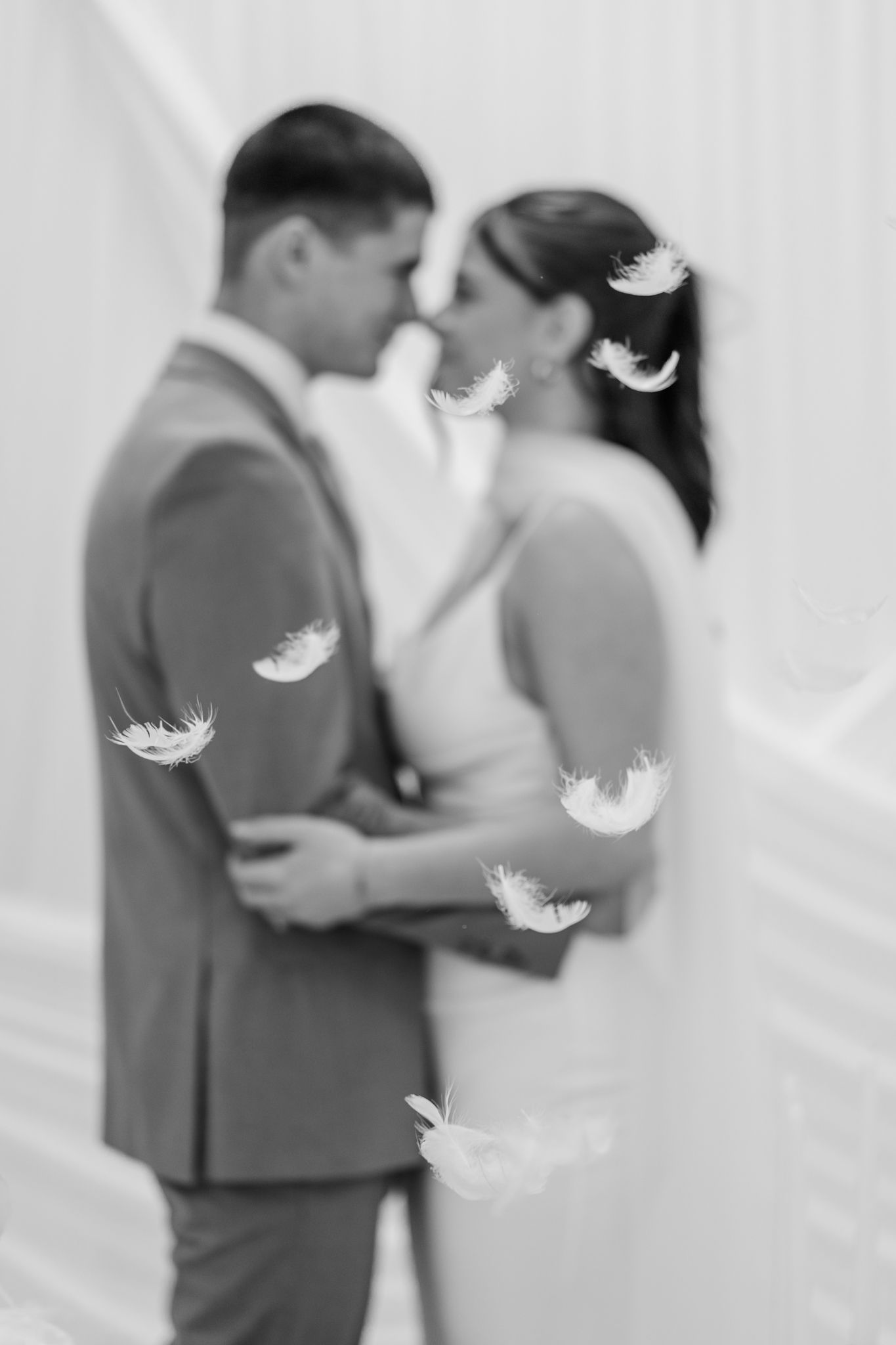 Black and white wedding photo of couple almost kissing with white feathers floating around them.
