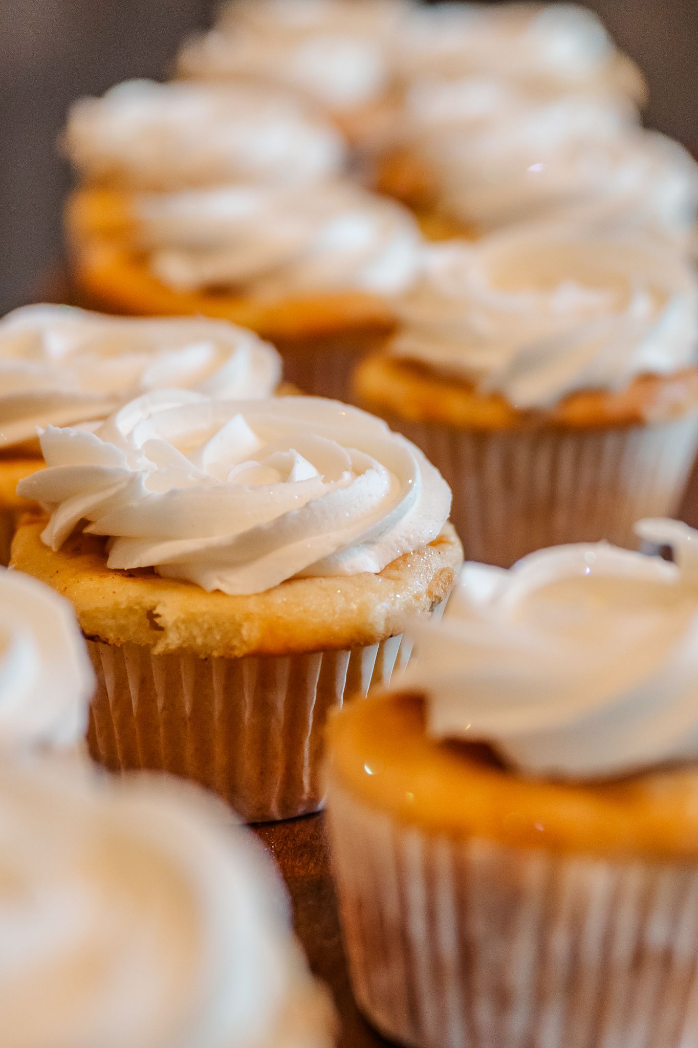 Rows of golden vanilla cupcakes topped with swirled white frosting displayed on a wooden surface.