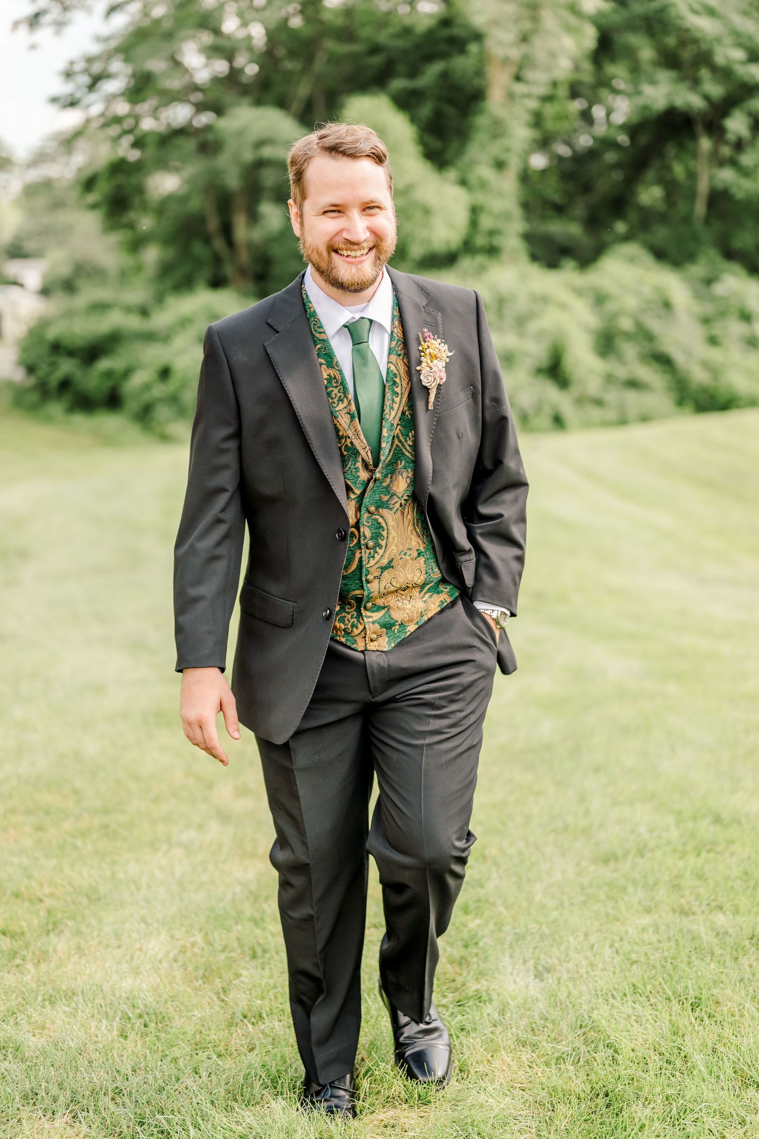 Wedding party member in black suit with green floral vest walking up grassy hill.
