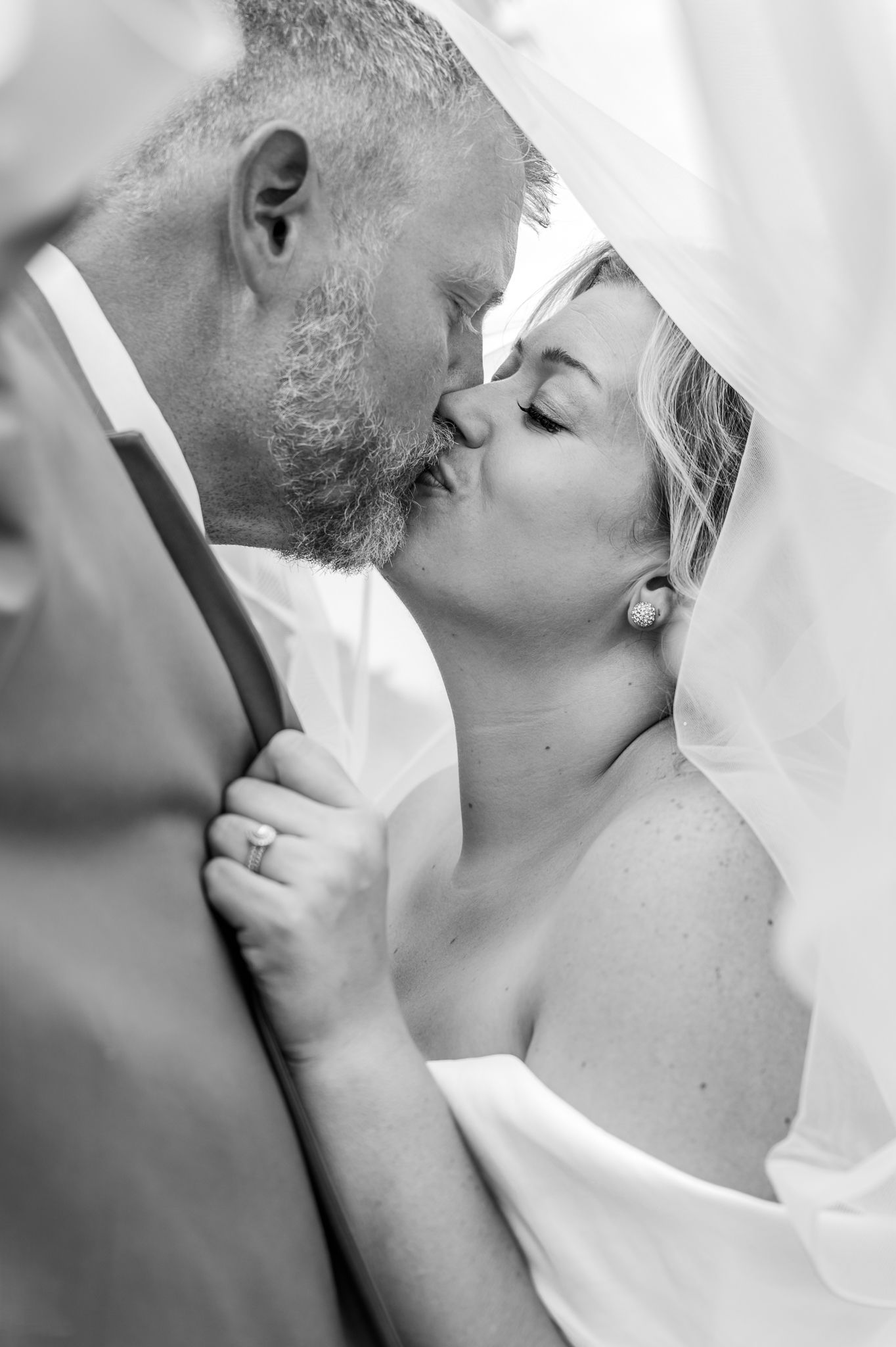 Black and white photo of a bride and groom sharing a tender kiss under a flowing veil on their wedding day. Genesis Grove Lodging and Events