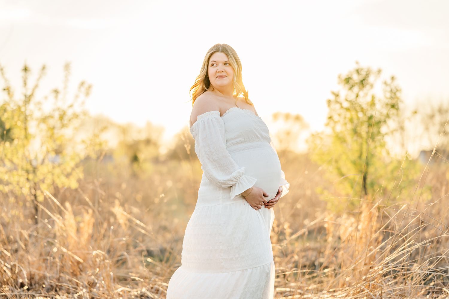 Pregnant woman in white off-shoulder dress standing in golden field at sunset, cradling baby bump.