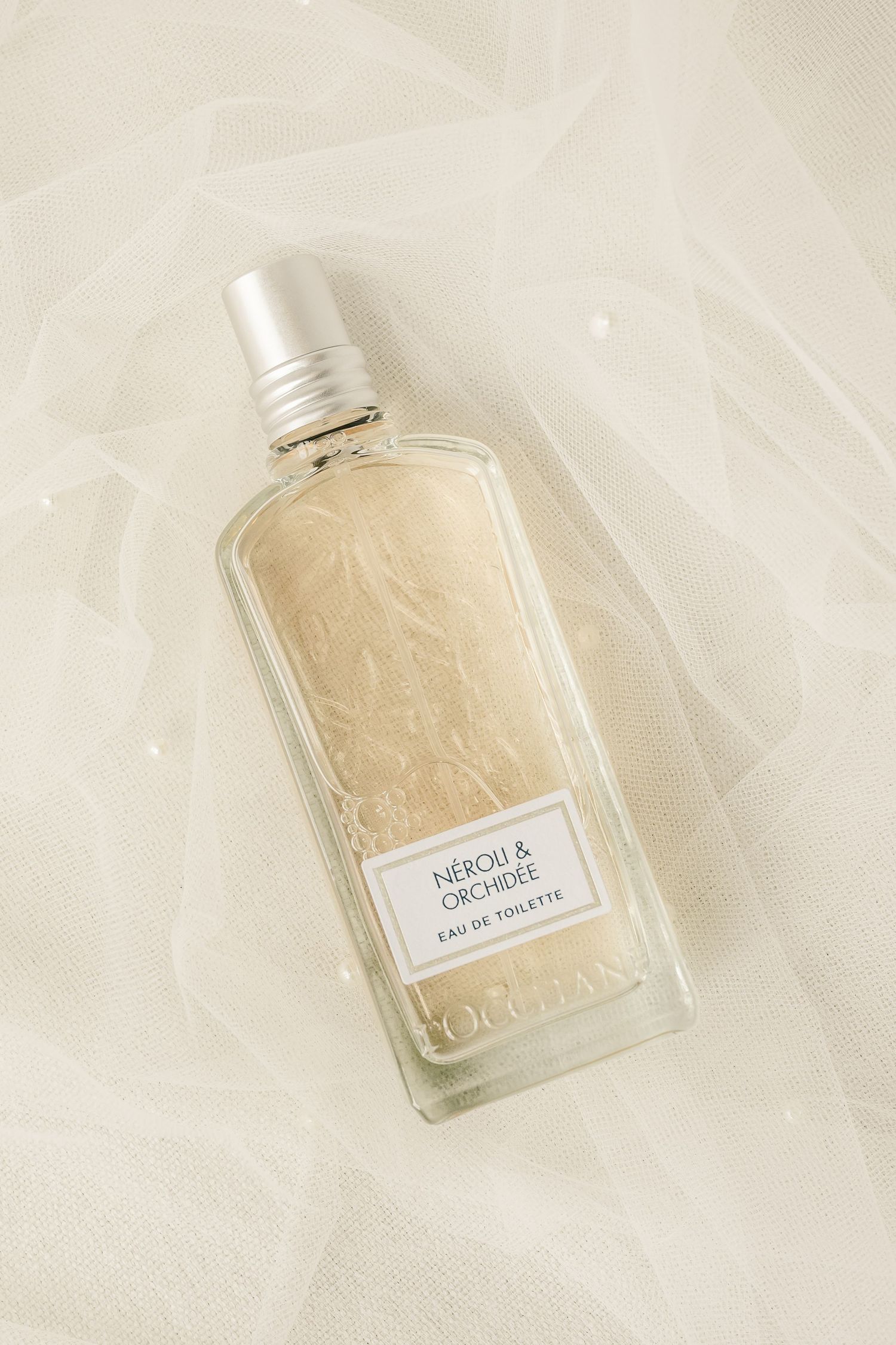 Elegant perfume bottle with beige liquid photographed on white silk fabric in soft natural lighting.