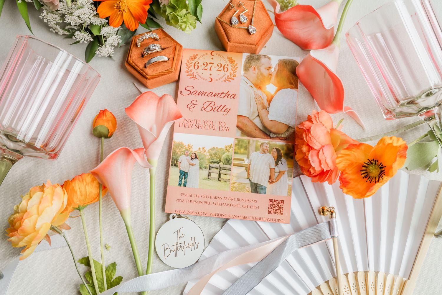 Elegant wedding flat lay with coral calla lilies, orange poppies, save-the-date card, rings, and decorative fan on white surface.