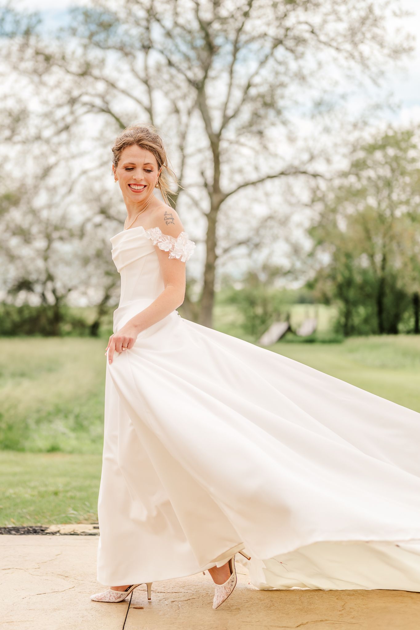Smiling bride in off-shoulder white gown twirling outdoors on stone patio with blooming trees in background.The Old Barns at Dry Run Farms.