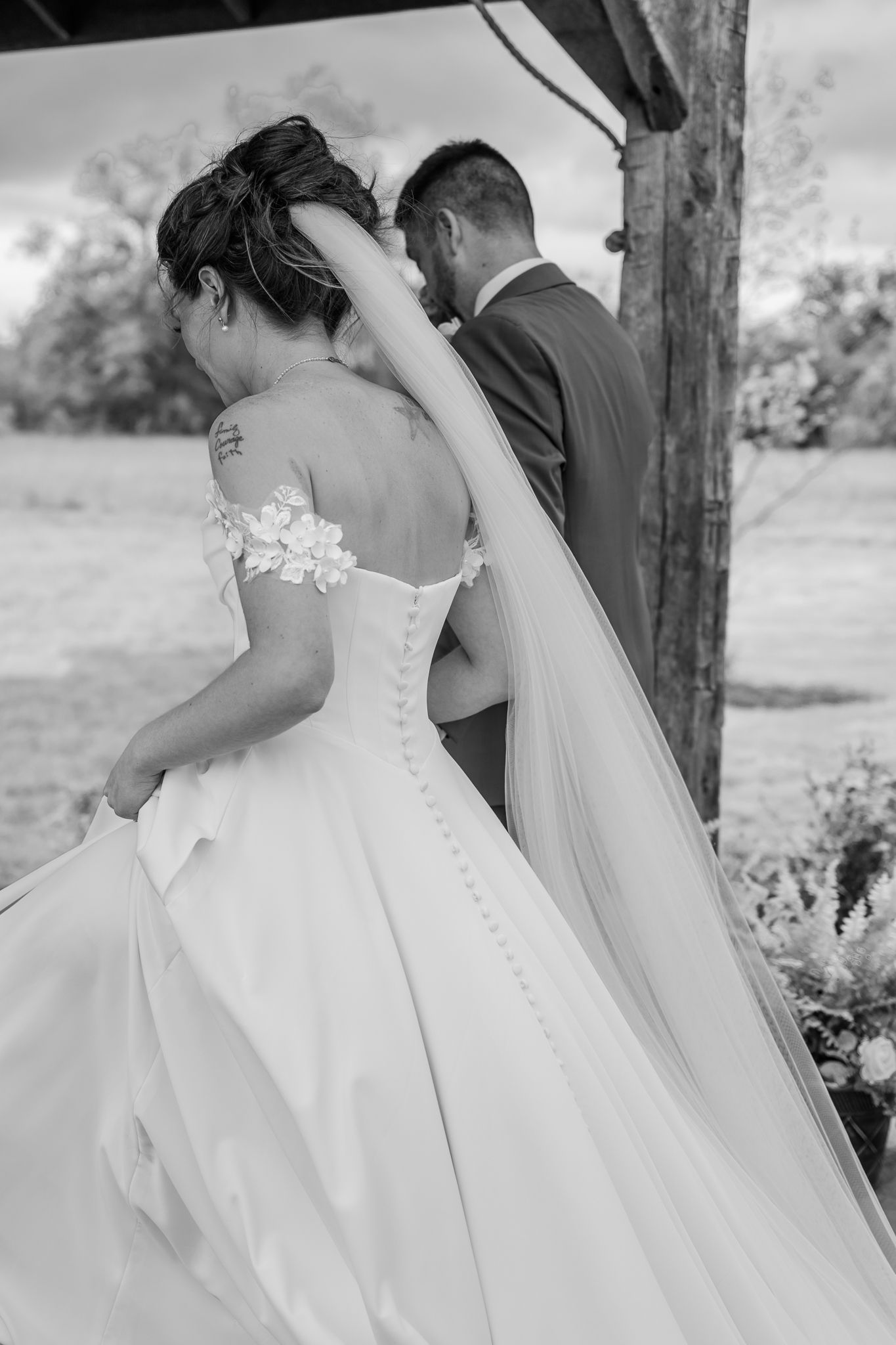 Black and white photo of bride in off-shoulder gown with veil and groom in suit standing under outdoor wedding altar.