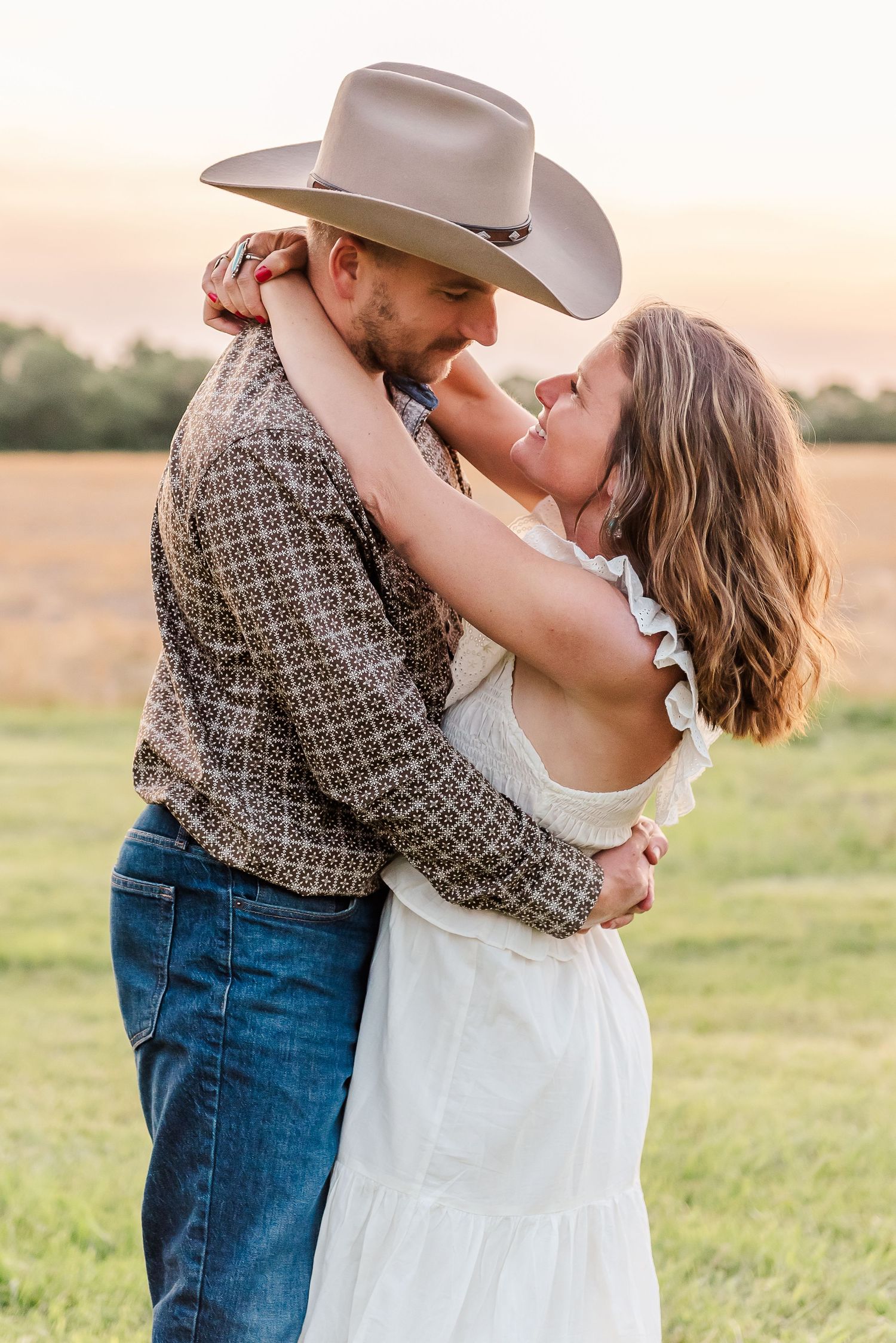 A couple in western attire embraces romantically in a sunlit field during golden hour.