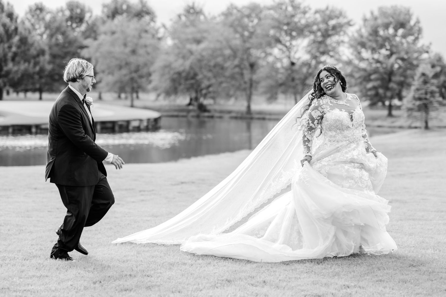 Black and white wedding photo of a bride in a flowing gown and long veil dancing joyfully with a groom in a suit on a lawn by a lake.
