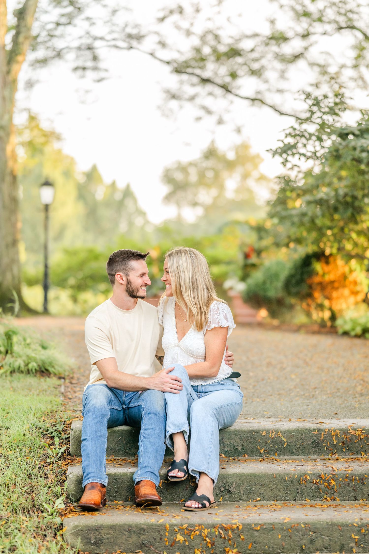 Smiling couple sits close together on stone steps in a sunlit park surrounded by autumn foliage during an outdoor engagement photo session.