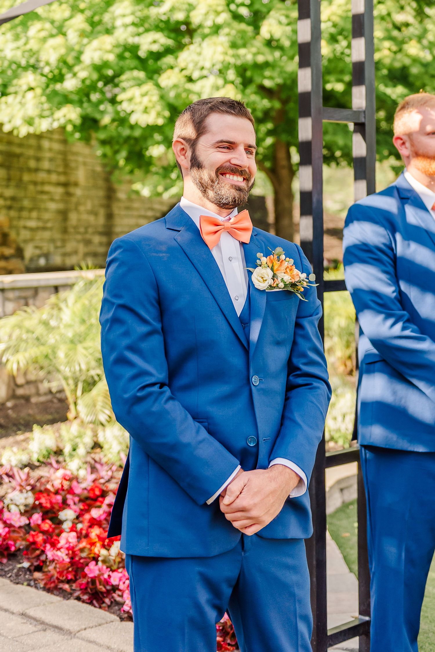 Groom in bright blue suit with coral bow tie and floral boutonniere stands smiling at outdoor wedding ceremony.