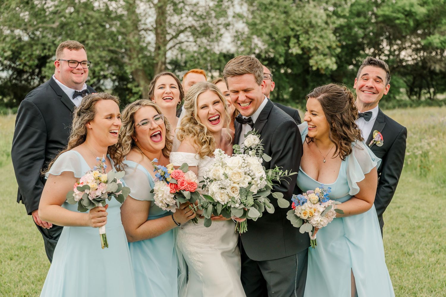 Wedding party laughs joyfully together while posing outdoors in light blue bridesmaid dresses and formal attire.