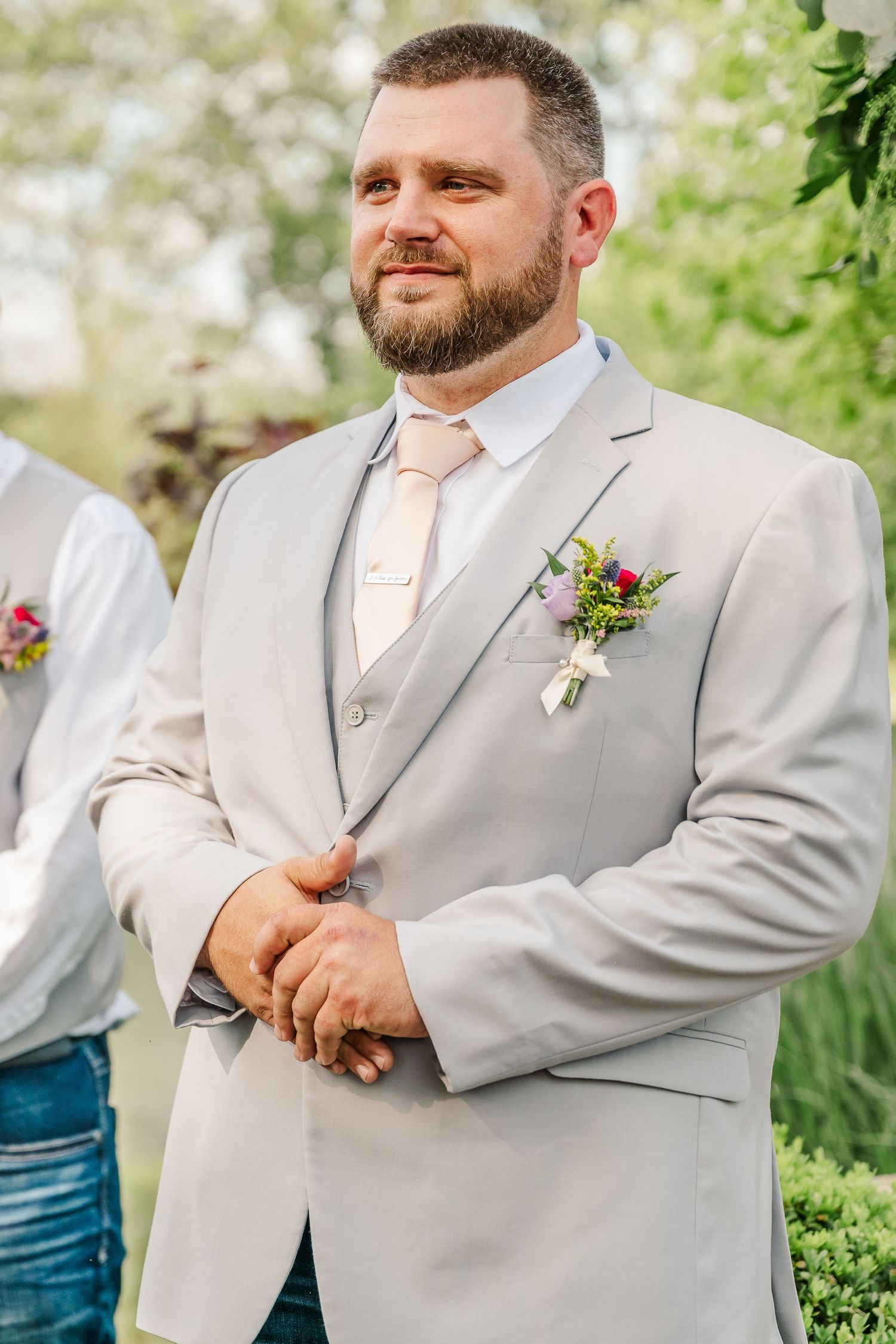 A groom in a light gray suit with a peach tie and boutonniere poses in a garden setting.