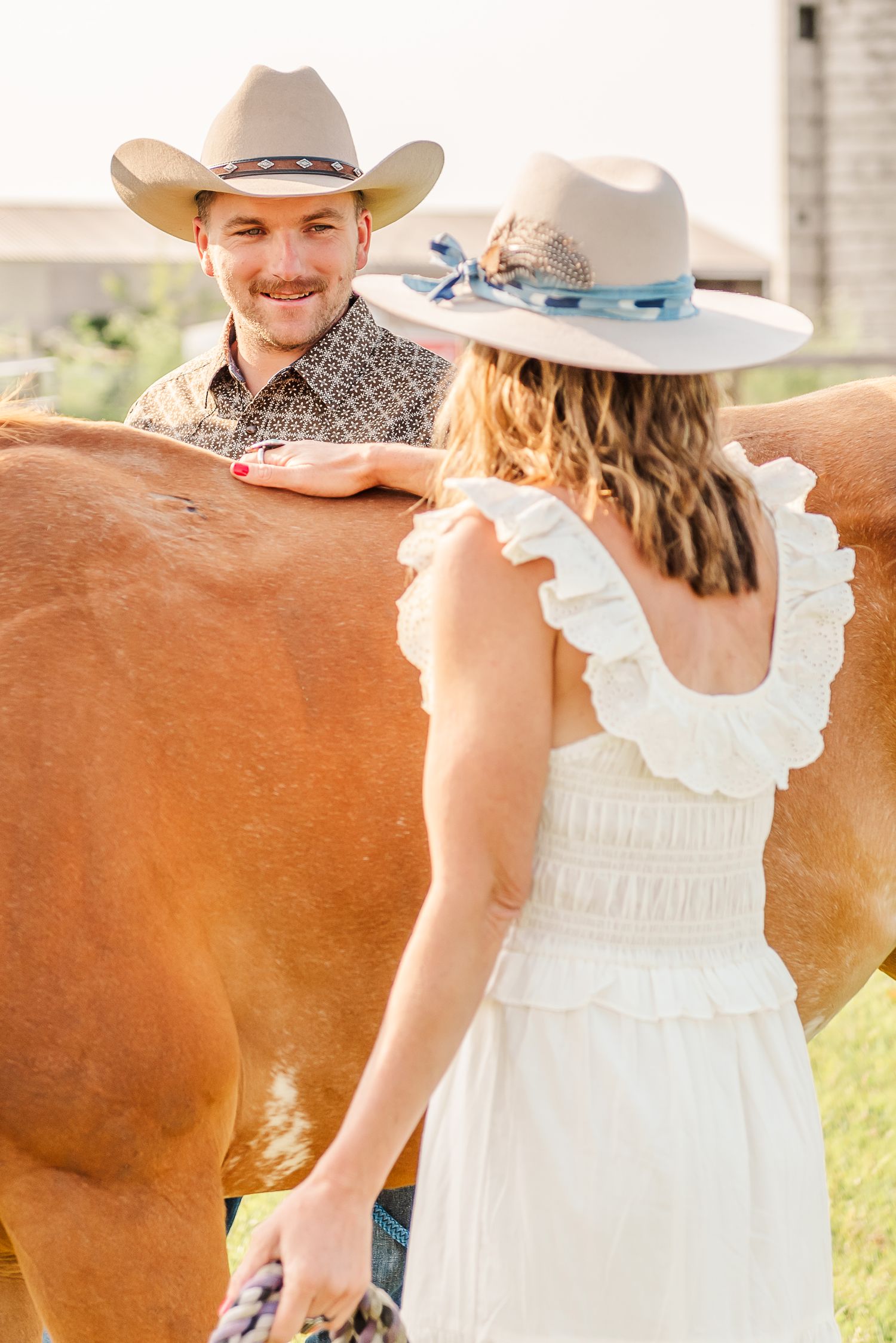 A couple in western attire poses with a brown horse in a rustic outdoor setting on a sunny day.