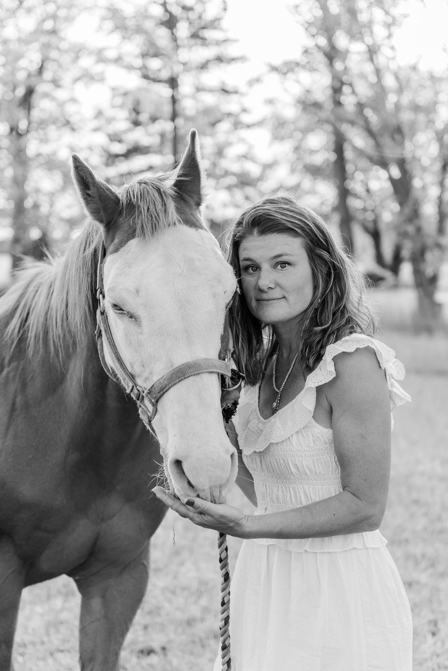 A woman in a white dress poses with a beautiful white horse in a dreamy outdoor setting.