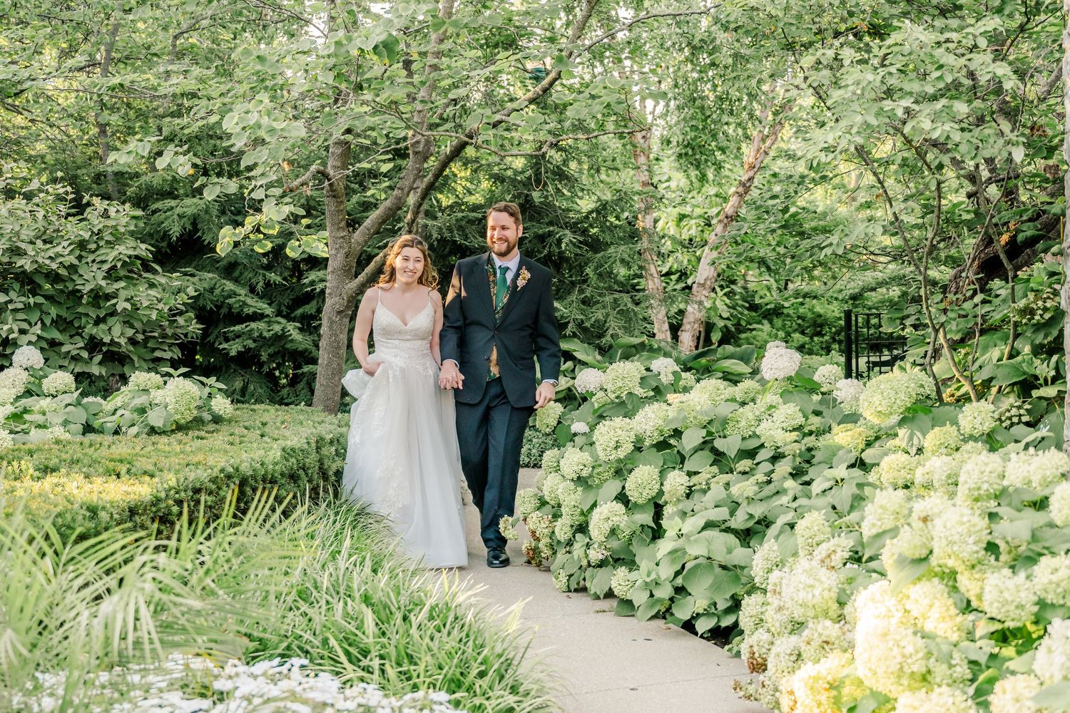 A couple walks together through a lush garden path surrounded by vibrant greenery and blooming flowers.