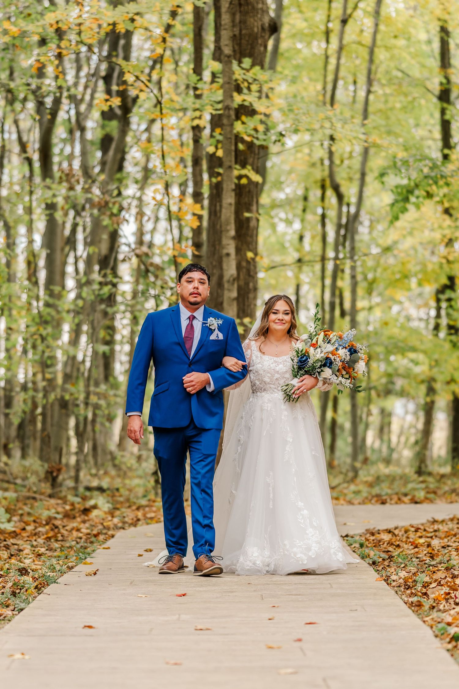 Wedding couple walks along tree-lined autumn path in coordinated navy and white attire. Four Seasons Barn Wedding & Event Venue