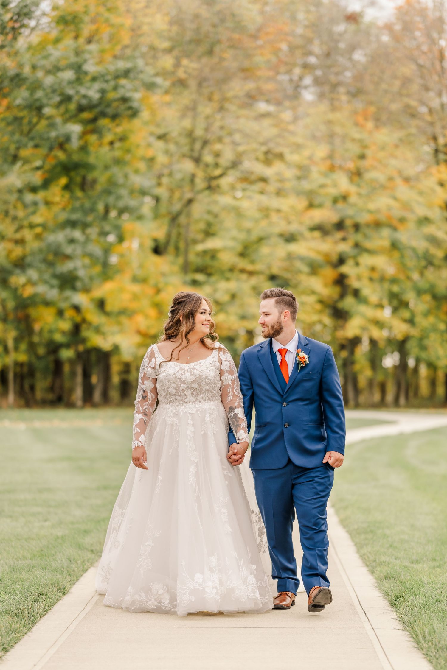 A couple walks hand in hand along a path surrounded by autumn trees with golden yellow foliage. Four Seasons Barn Wedding & Event Venue