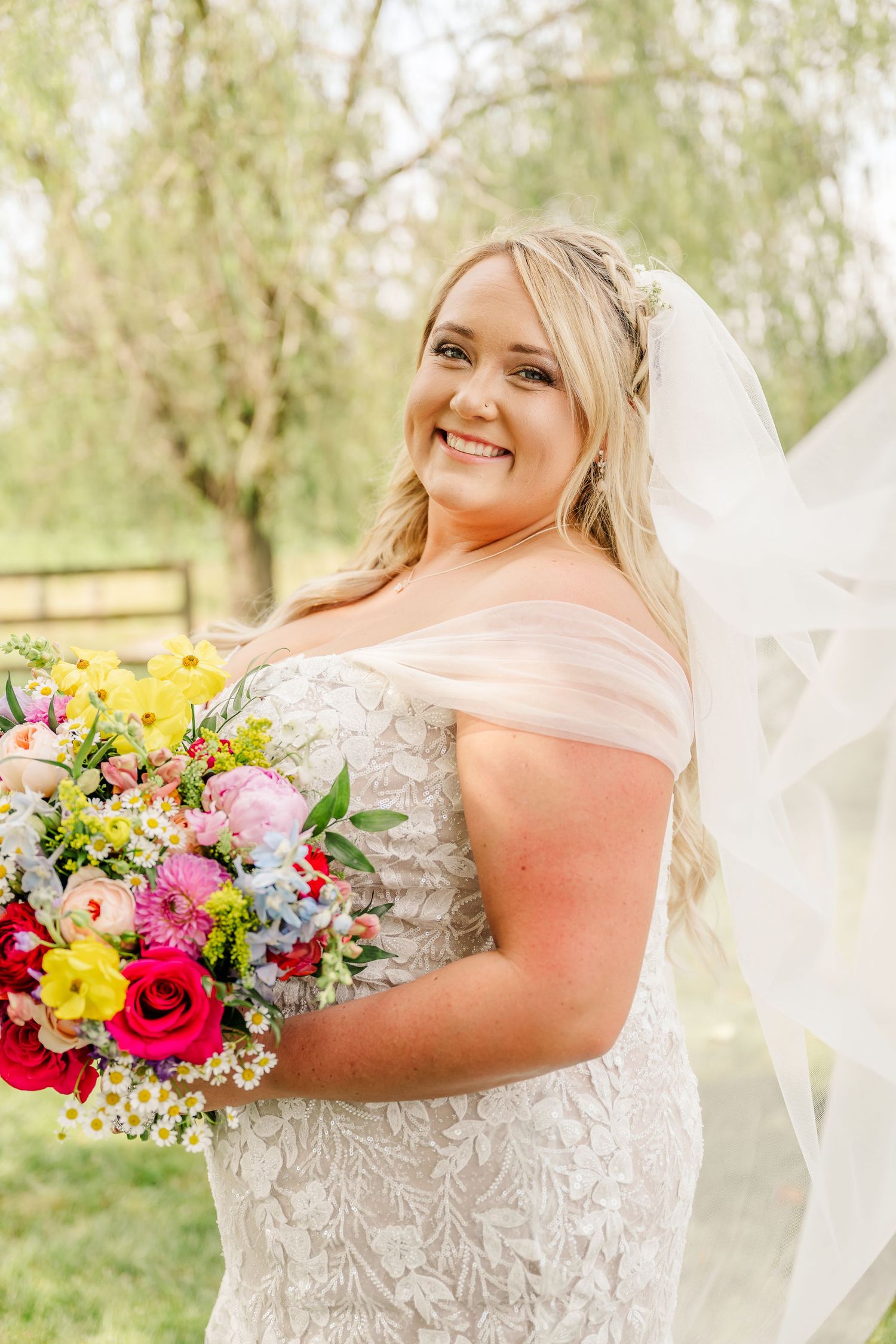 A bride in an off-shoulder lace gown holds a vibrant bouquet of pink, yellow and red flowers against a garden backdrop.