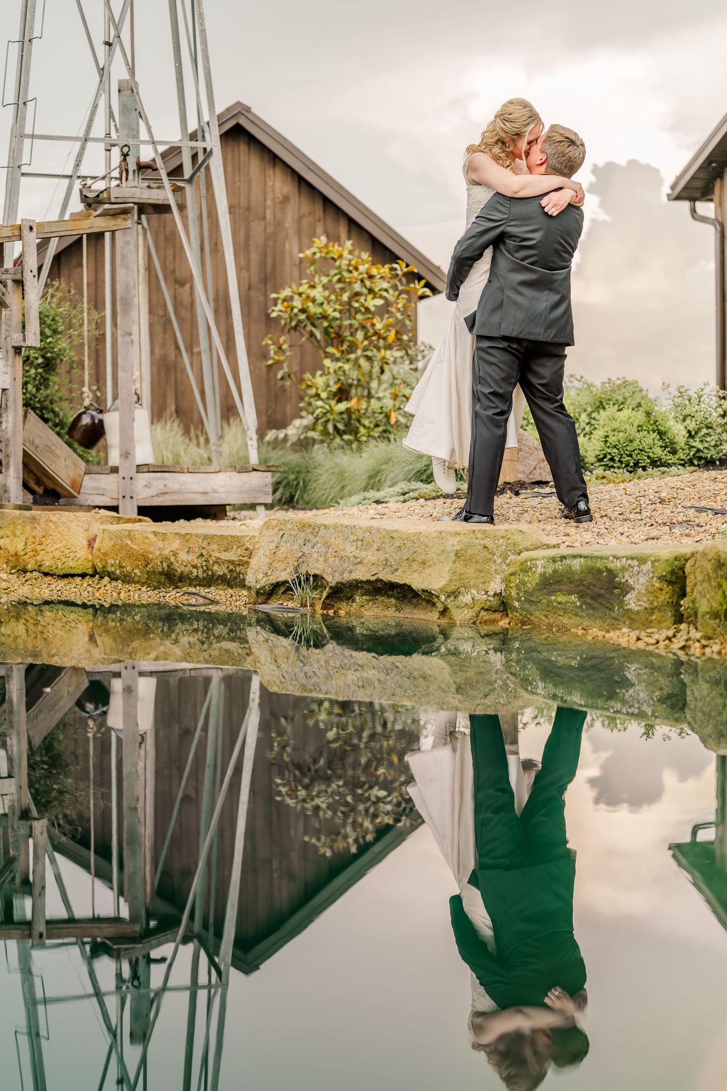 A romantic embrace on stone steps with a perfect reflection in still water below, creating a mirror image against a rustic barn.
