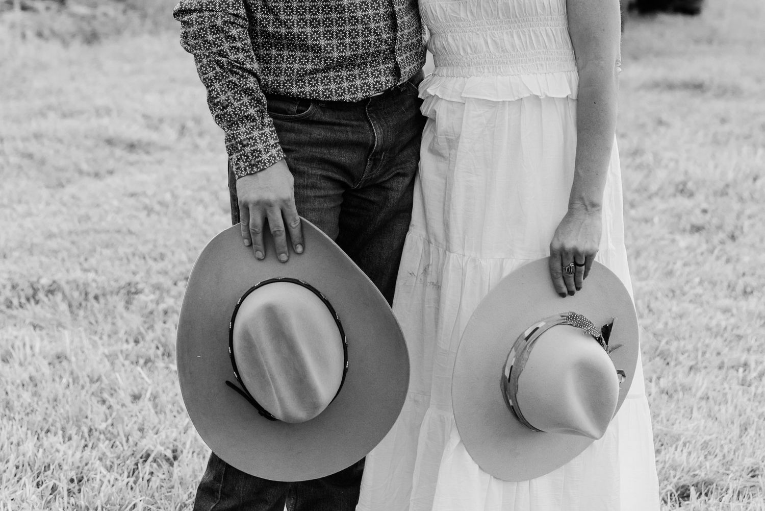 Black and white photo of a couple in western attire holding hands and cowboy hats while walking through a field.