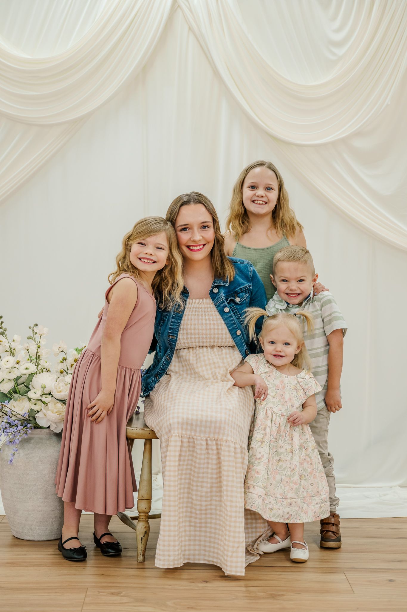 A young woman in a denim jacket sits with four children in front of white draping for a family portrait.