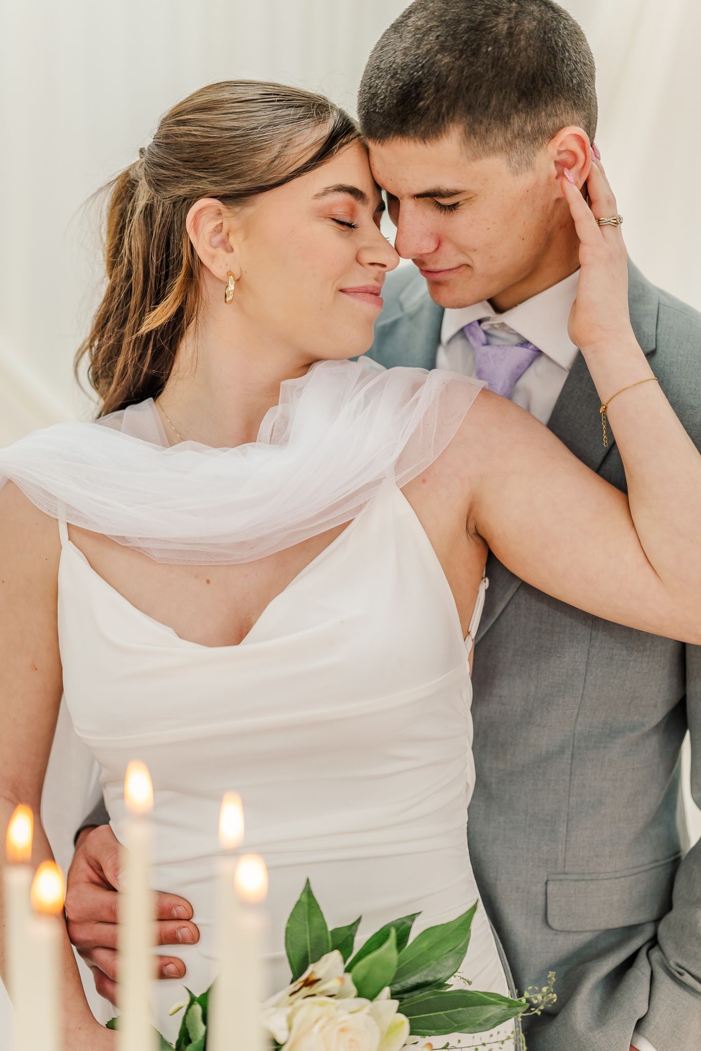 A bride and groom share an intimate moment, touching foreheads with candles and white flowers in the foreground.