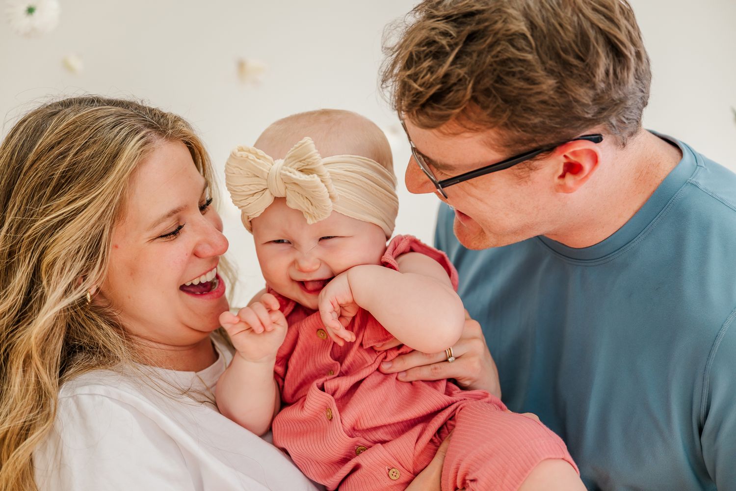 Happy family with parents laughing and smiling with their baby girl in a pink outfit and bow headband.