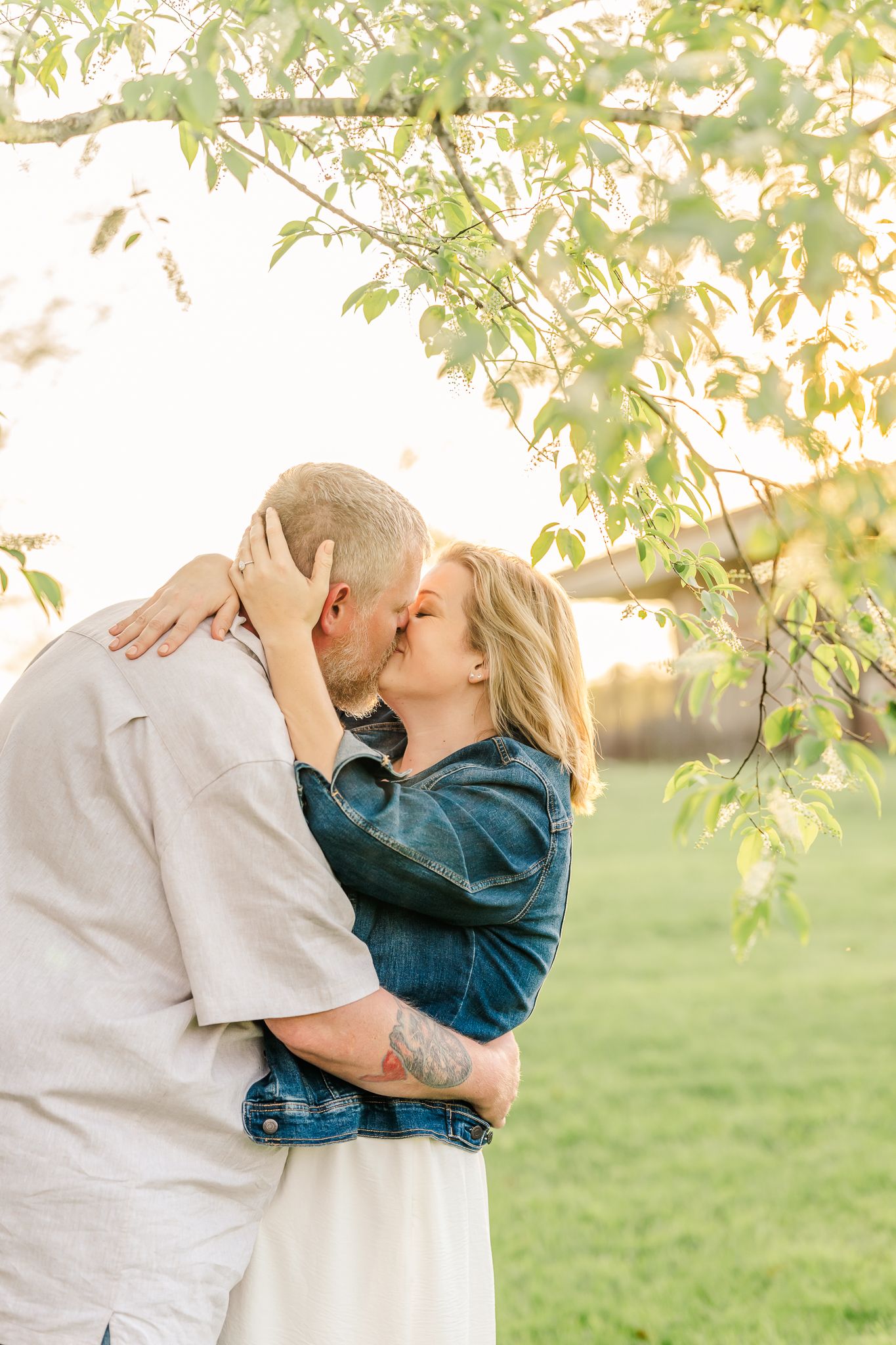 Couple sharing a romantic kiss under green tree branches in a sunlit outdoor setting, woman in denim jacket embracing man.