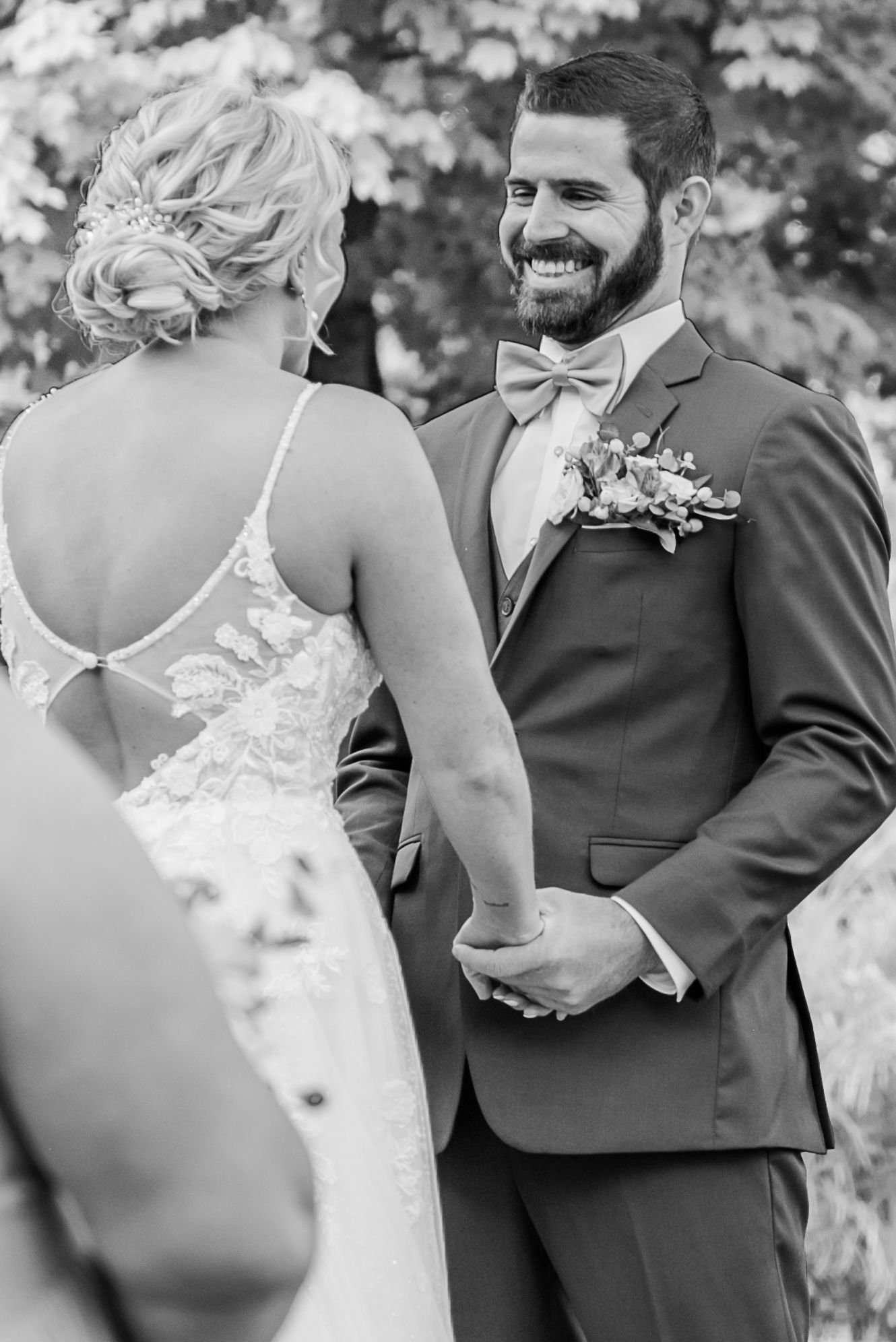 A black and white wedding photo captures a joyful moment between newlyweds as they hold hands outdoors.