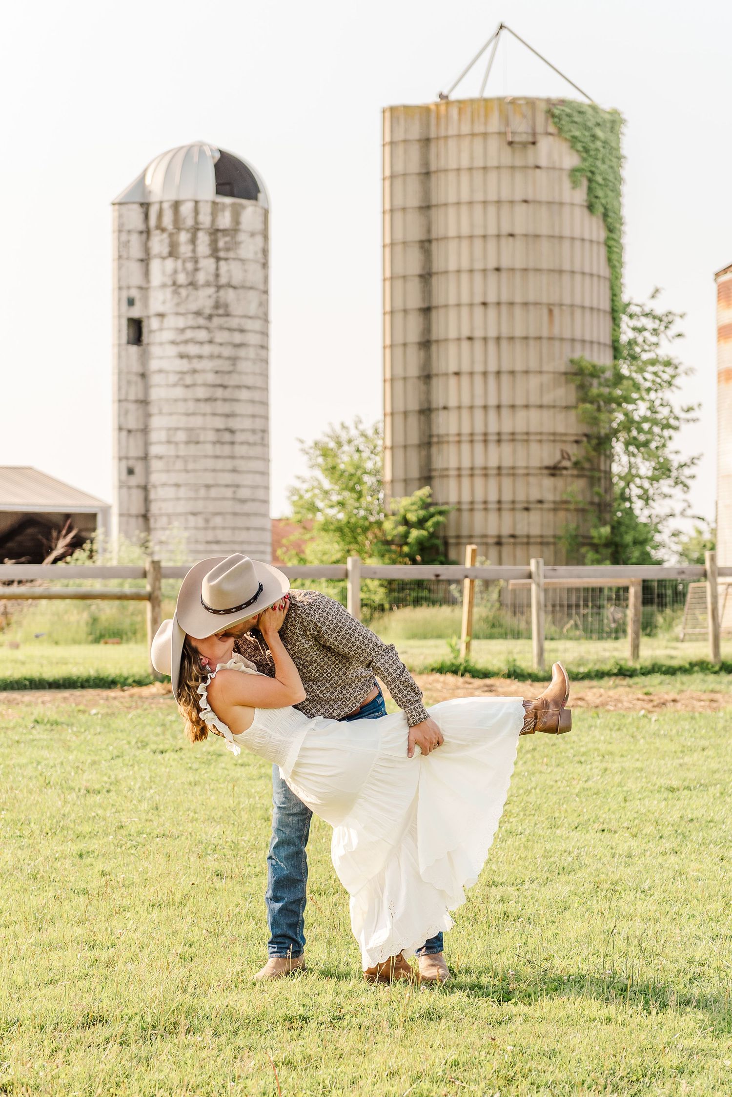 A romantic couple dips and dances together in a field with rustic grain silos in the background.