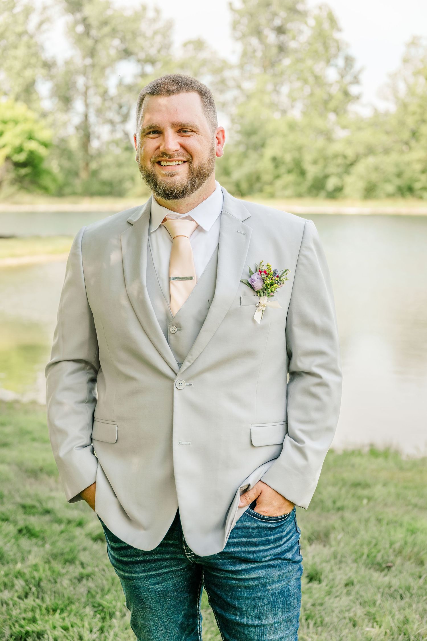 A person in a light gray suit jacket and jeans stands smiling by a lake on a sunny day.