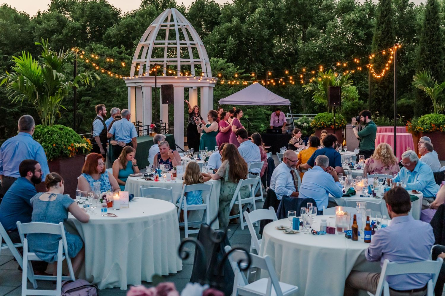 Elegant outdoor wedding reception with round tables and string lights under a garden gazebo.
