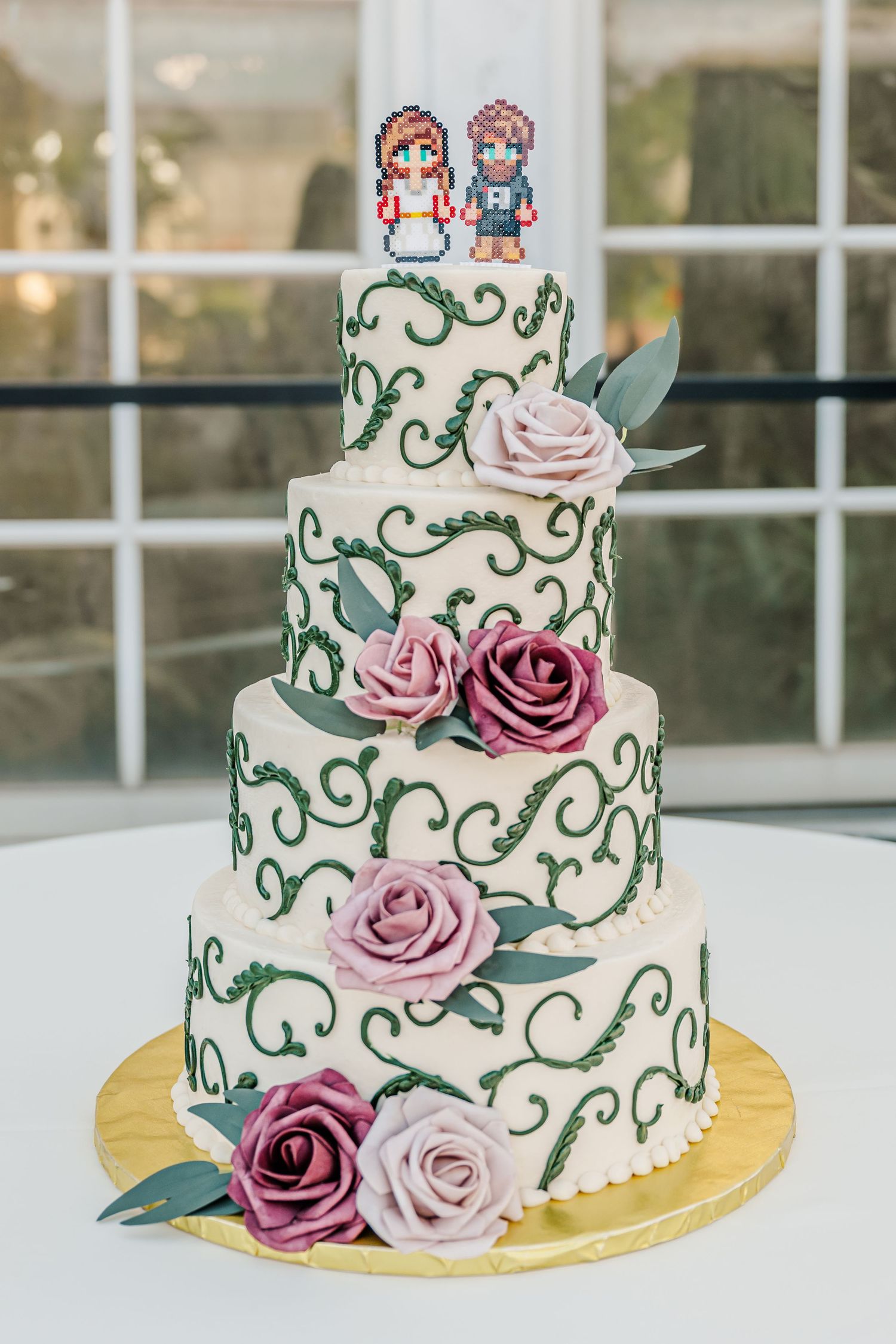 Three-tiered white wedding cake decorated with green swirls, pink and purple roses, and pixel art cake toppers.
