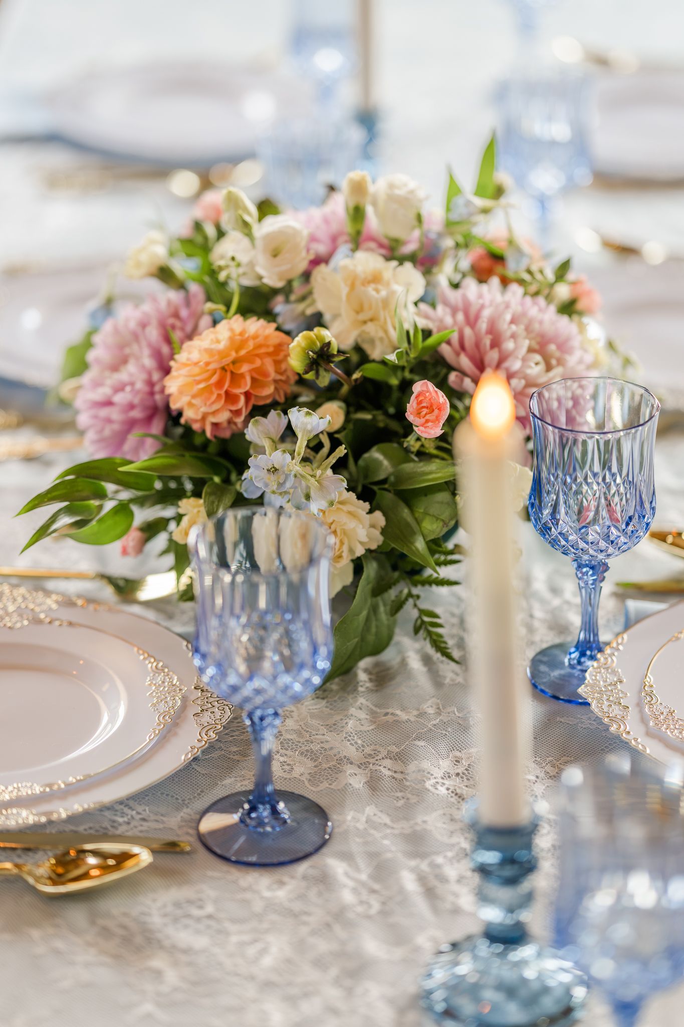 Elegant wedding table setting with blue crystal glasses, lit candle, gold accents, and a floral centerpiece of pink and white blooms.