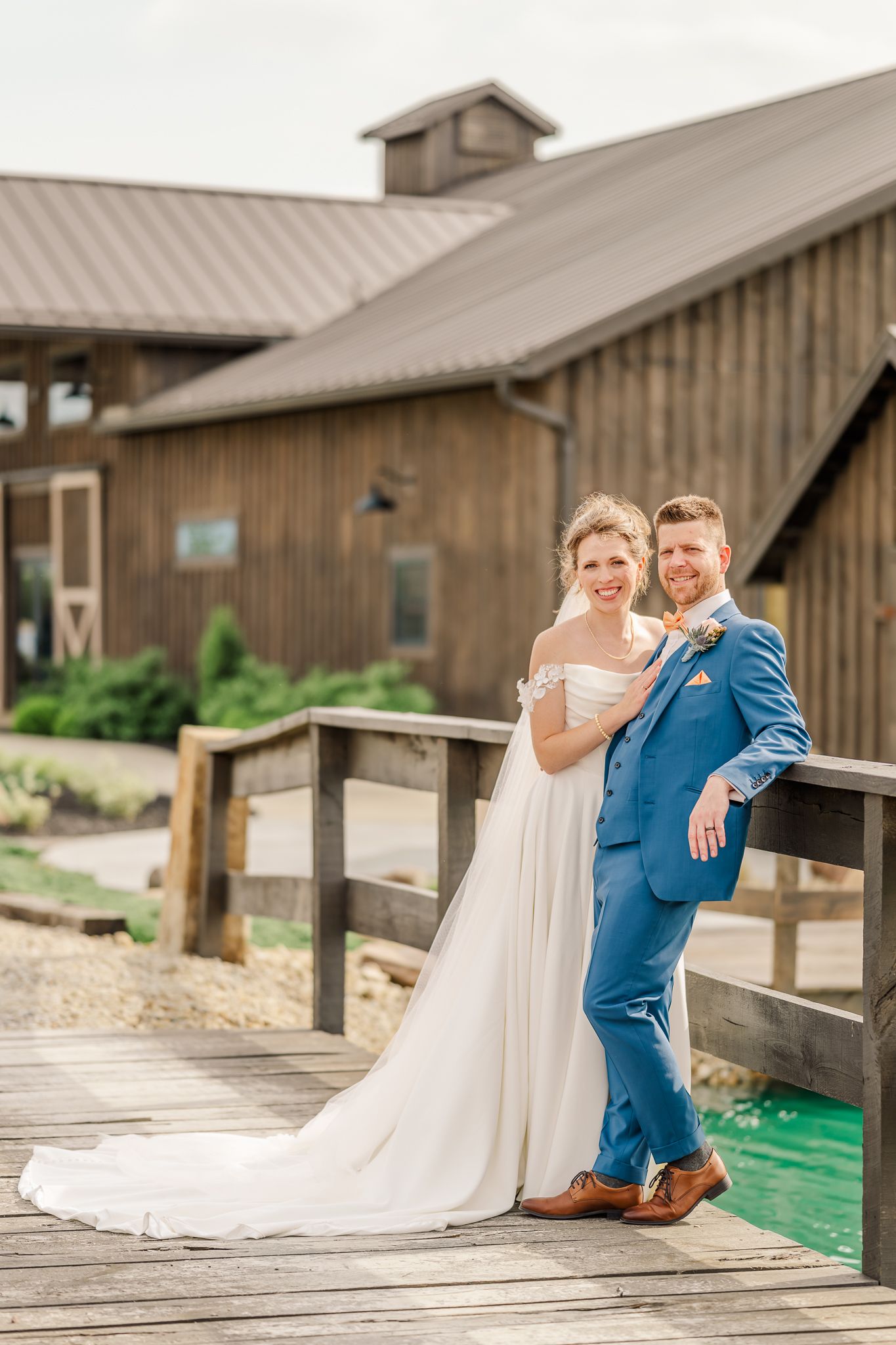 Bride and groom posing together on a wooden bridge in front of a rustic barn venue. The Old Barns at Dry Run Farms in Williamsport, Ohio.